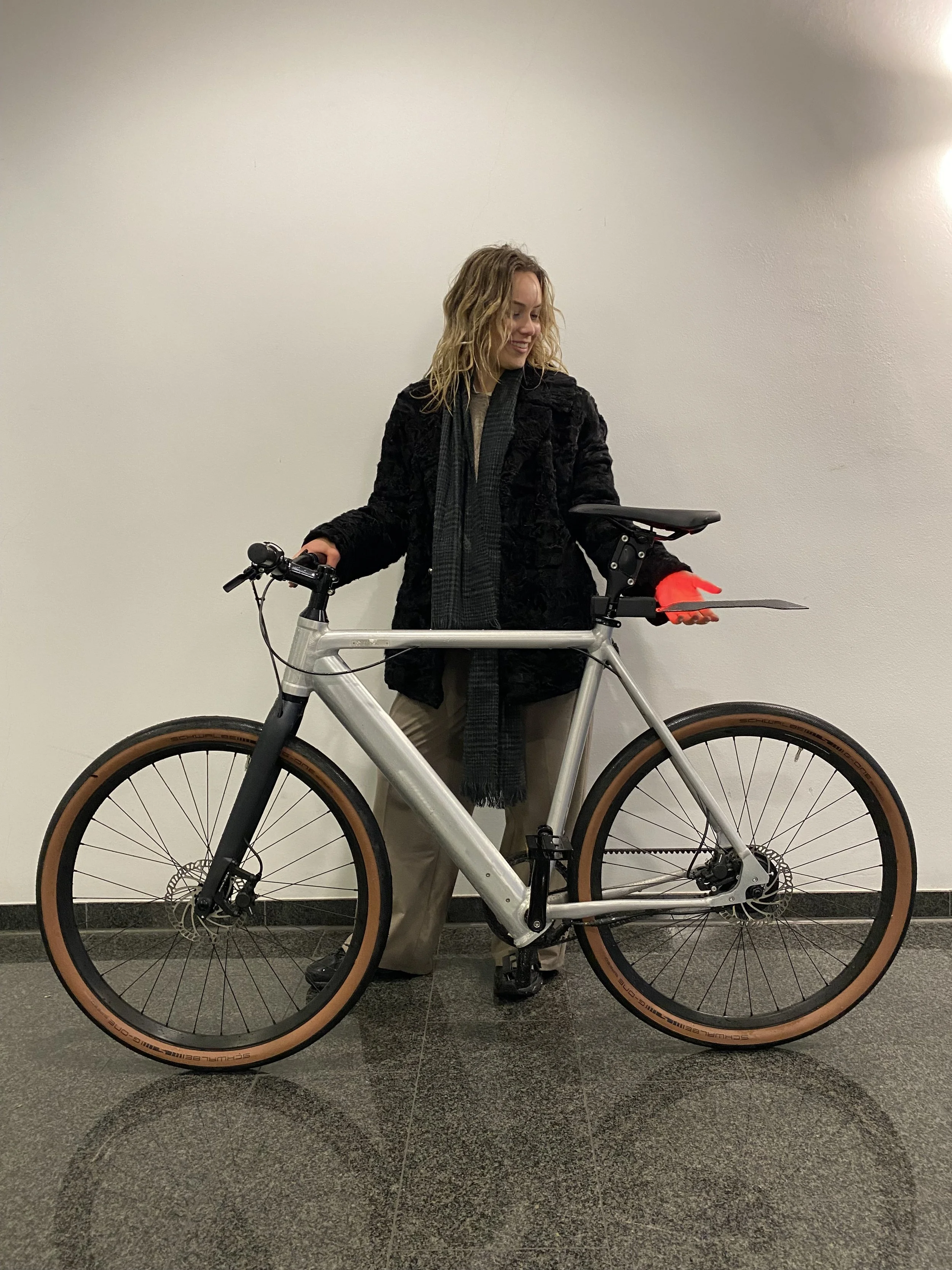 Person standing inside holding a bicycle with a silver frame and brown tires, against a plain wall.