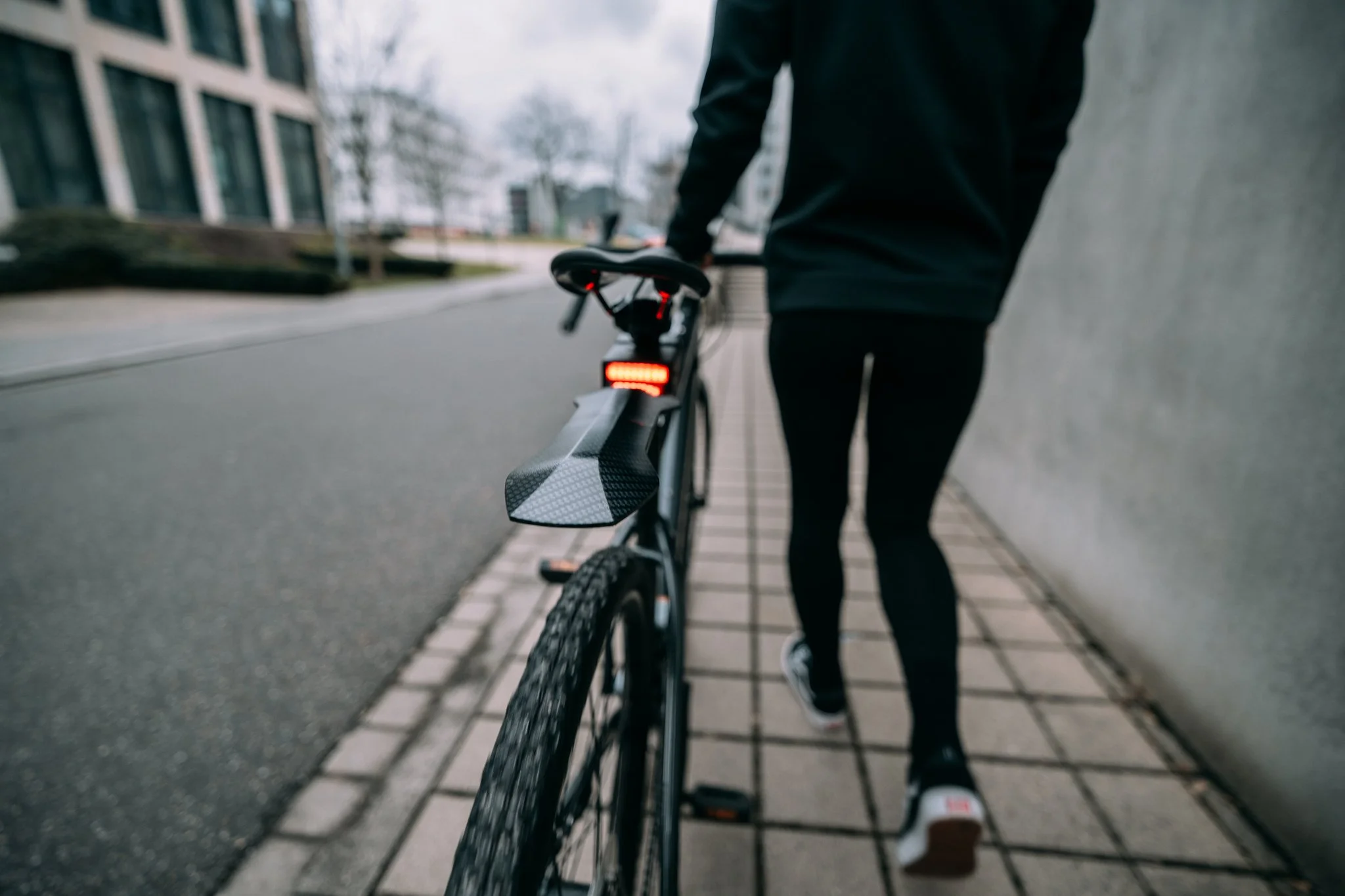 Person walking a bicycle on a city sidewalk, wearing black clothing, with a rear bike light on.