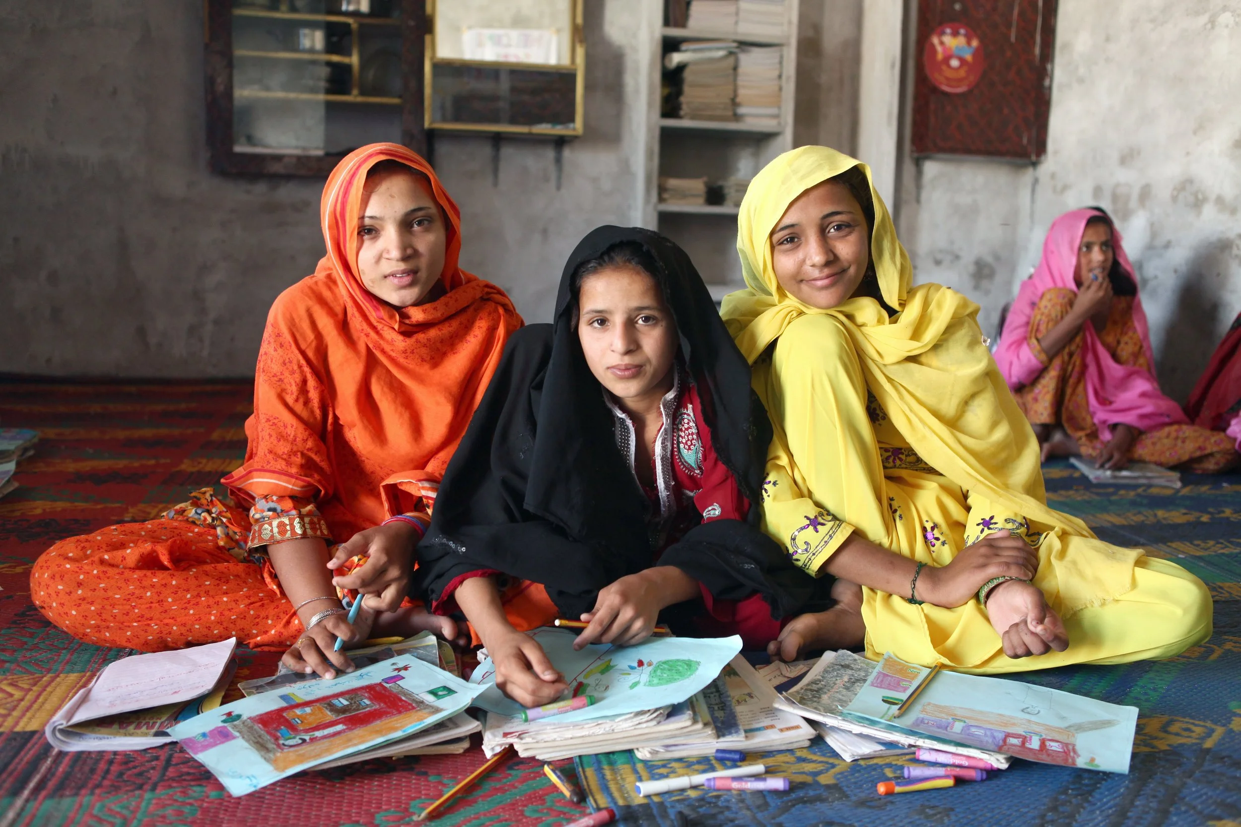 Three Pakistani school girls sitting on the floor behind textbooks, wearing orange, black, and yellow