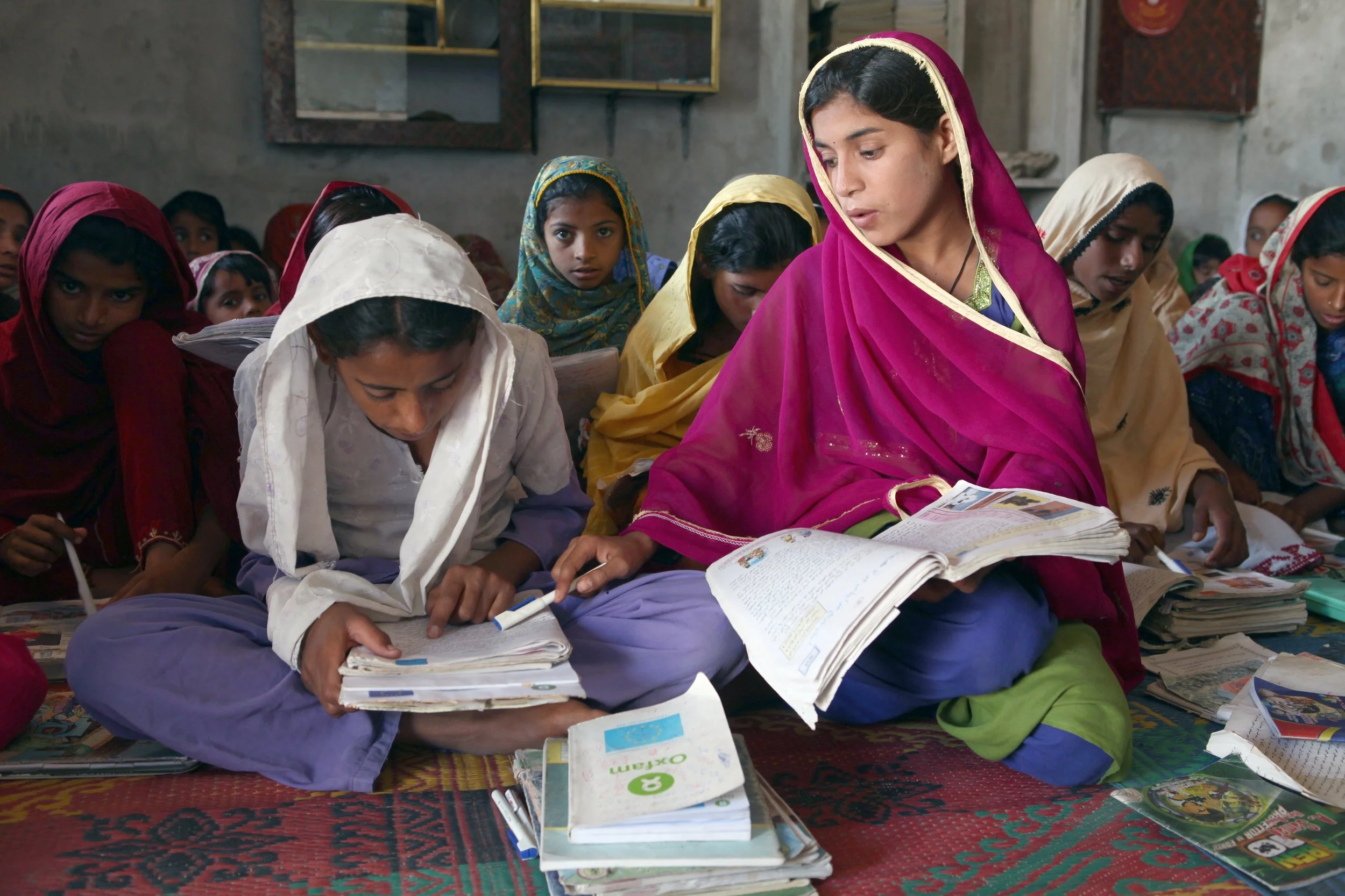 Pakistani school girls sitting cross legged on the floor and writing in textbooks, wearing colorful clothes