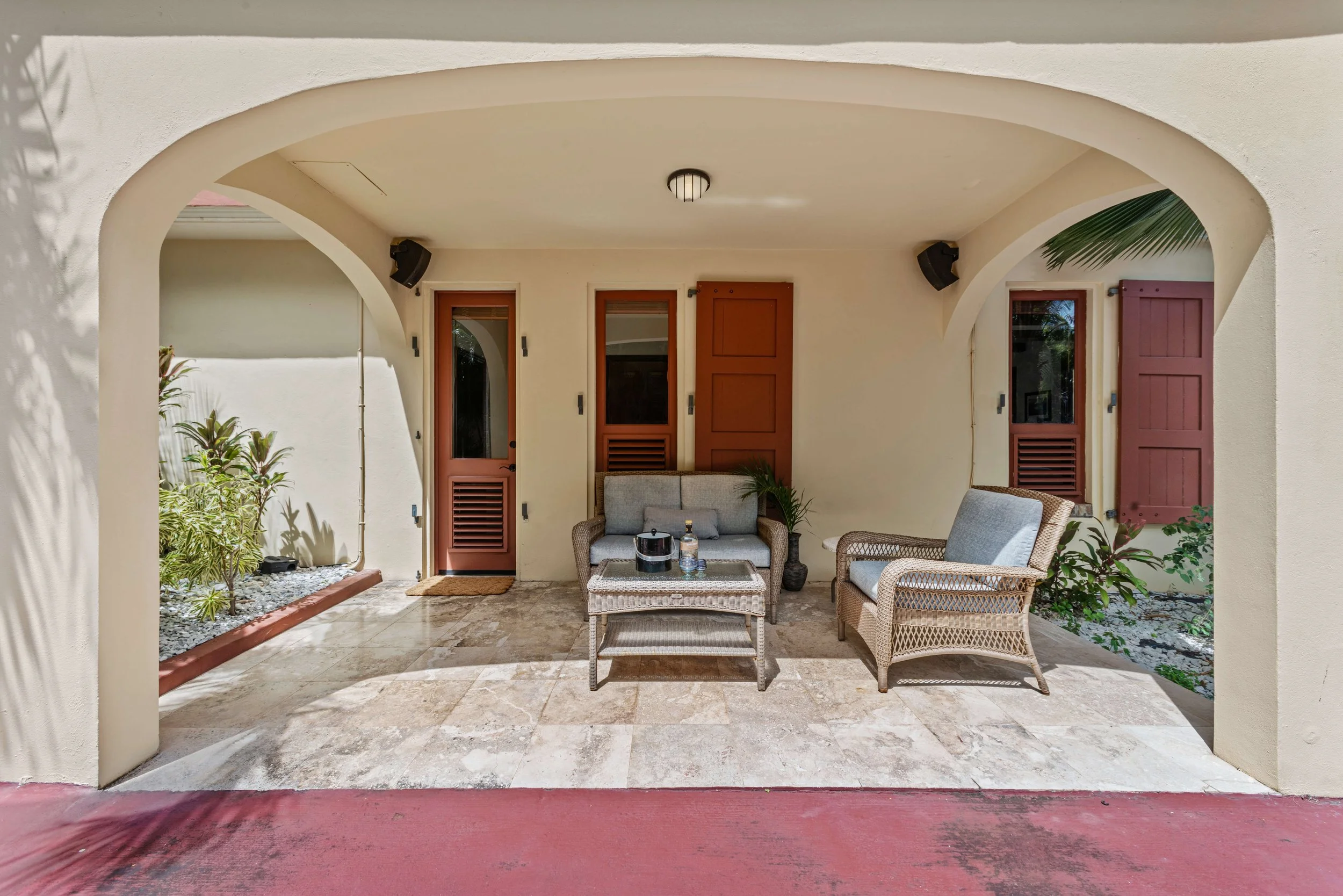 Covered patio with beige walls, stone flooring, wicker furniture including a loveseat, an armchair, and a coffee table. There are potted plants, brown doors with shutters, and a small plant on the right side.