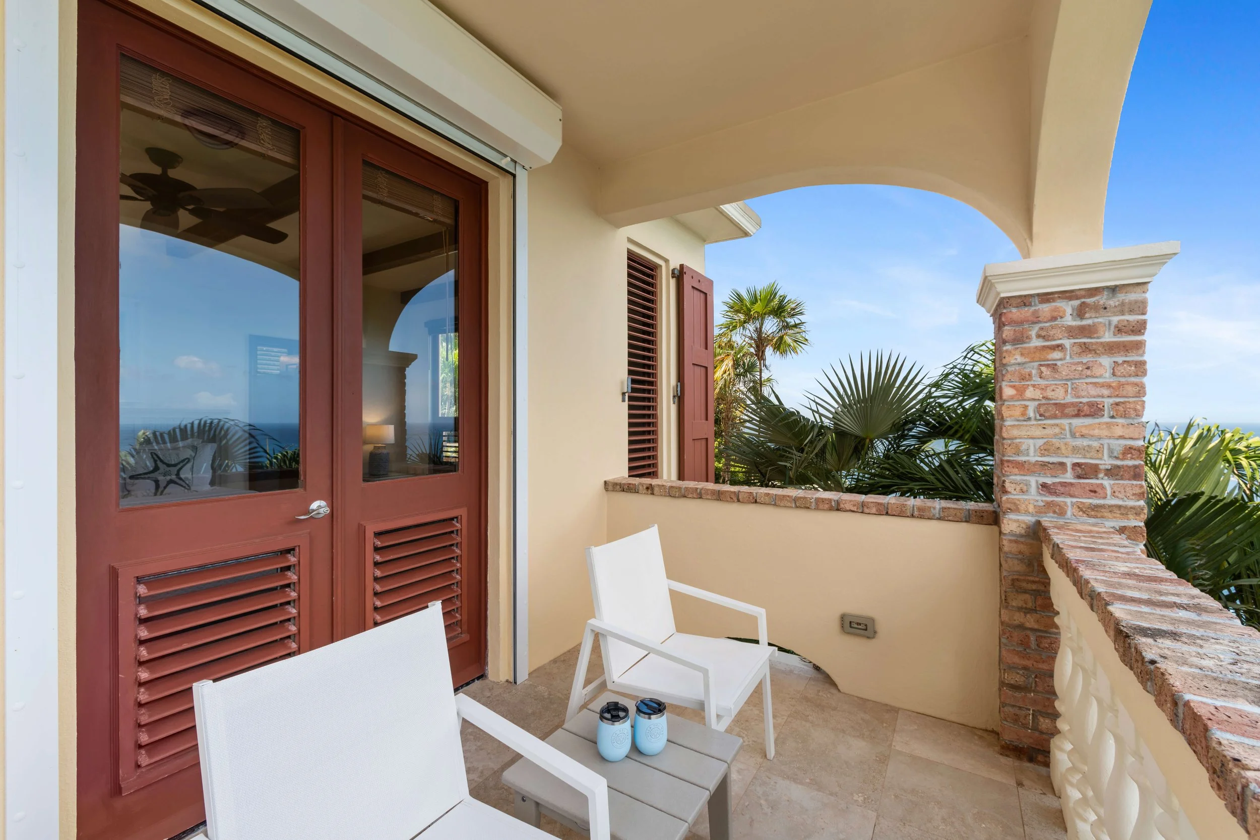 View of a balcony with two white chairs, a small table with two drinks, a brick railing, and tropical plants, overlooking a blue sky and ocean.