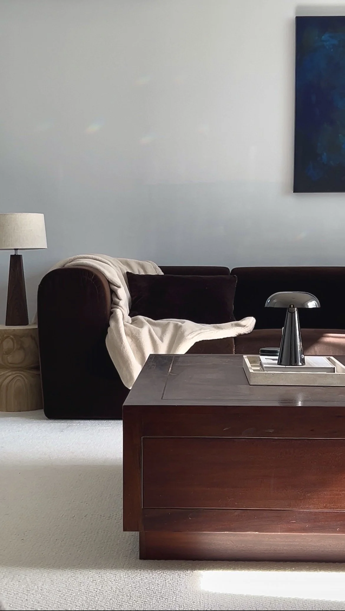 Contemporary living room with deep brown sofa, walnut table lamp with linen lampshade, dark timber coffee table, black chrome lamp on a tray, pale wool carpet, neutral throw, and dark blue artwork on a light grey wall.