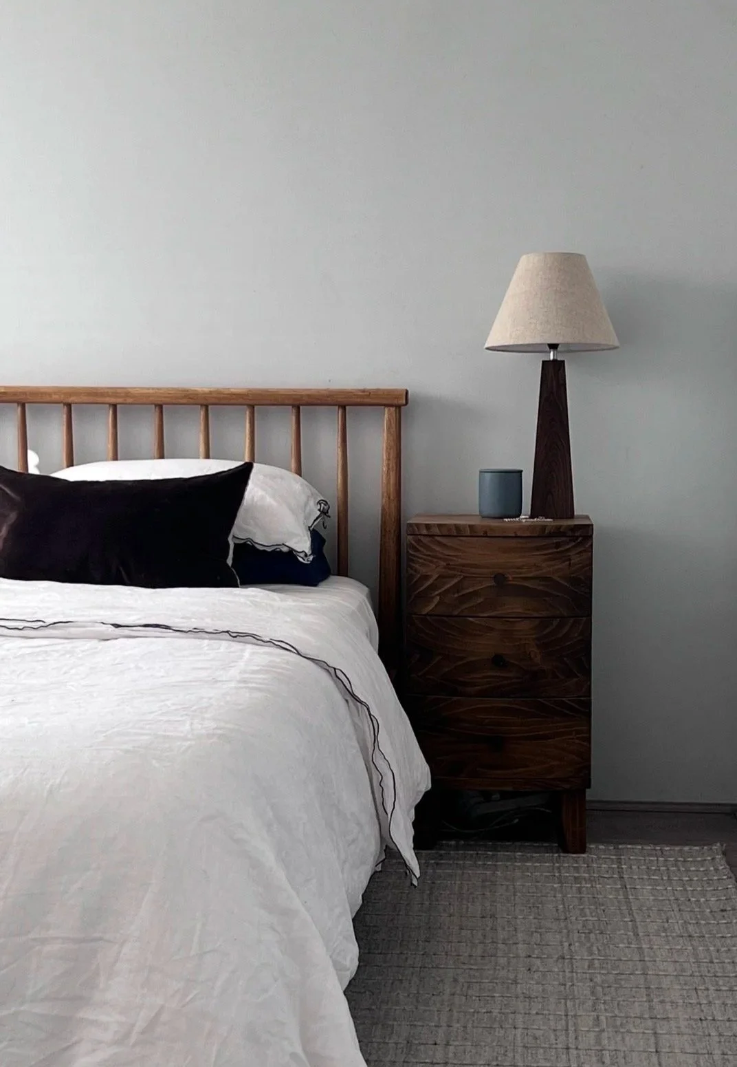 Contemporary bedroom with white linen bedding, timber bedhead, dark timber bedside table, walnut table lamp with linen lampshade, dark accent cushion, soft grey wall and textured neutral rug.