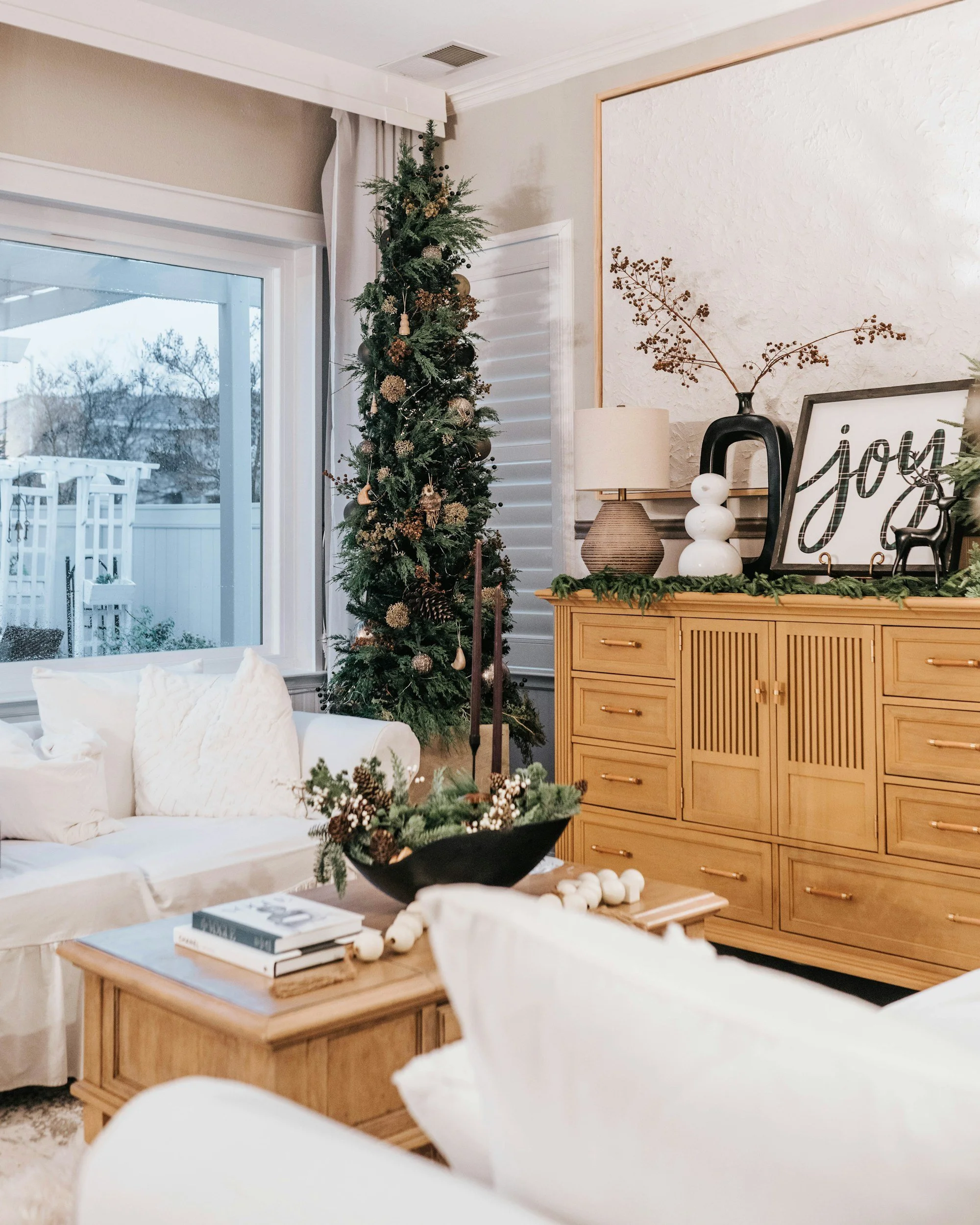 Living room with a tall Christmas tree decorated in neutral ornaments, next to a timber sideboard with greenery and sculptural decor.