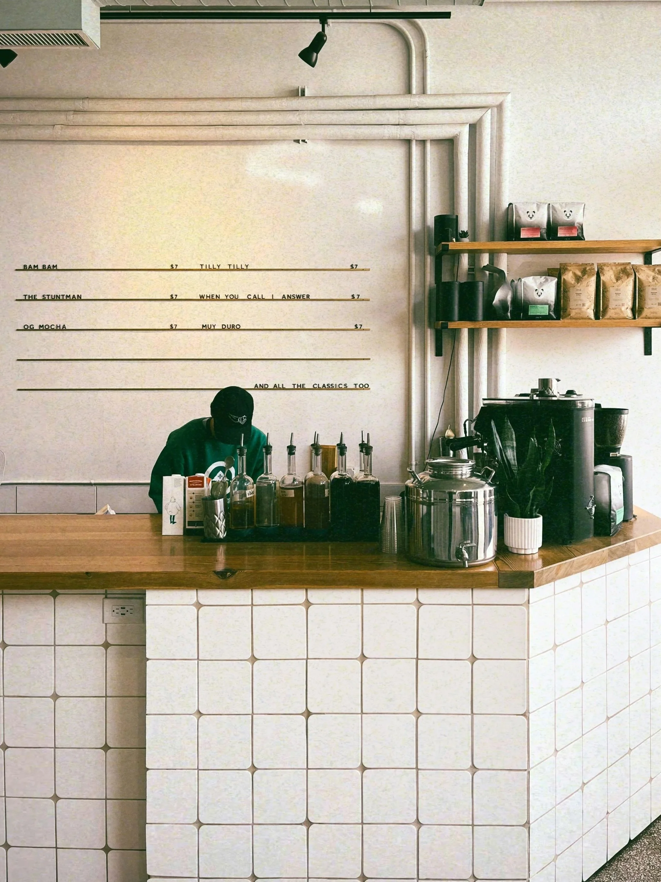 Interior of a coffee shop counter with a barista preparing drinks, coffee equipment, and shelves with coffee bags, against a tiled front and white wall with menu boards.