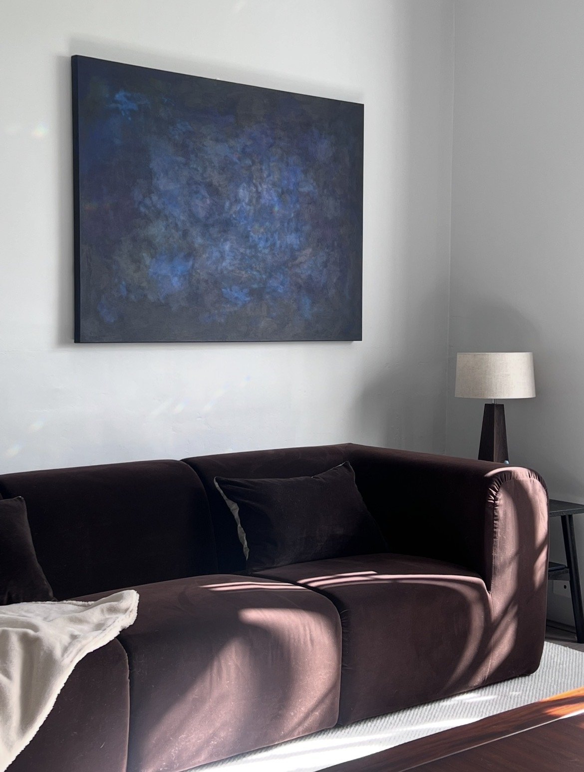 Contemporary living room with deep brown sofa, walnut table lamp with linen lampshade, dark timber coffee table, pale wool carpet, neutral throw, and dark blue artwork on a light grey wall.