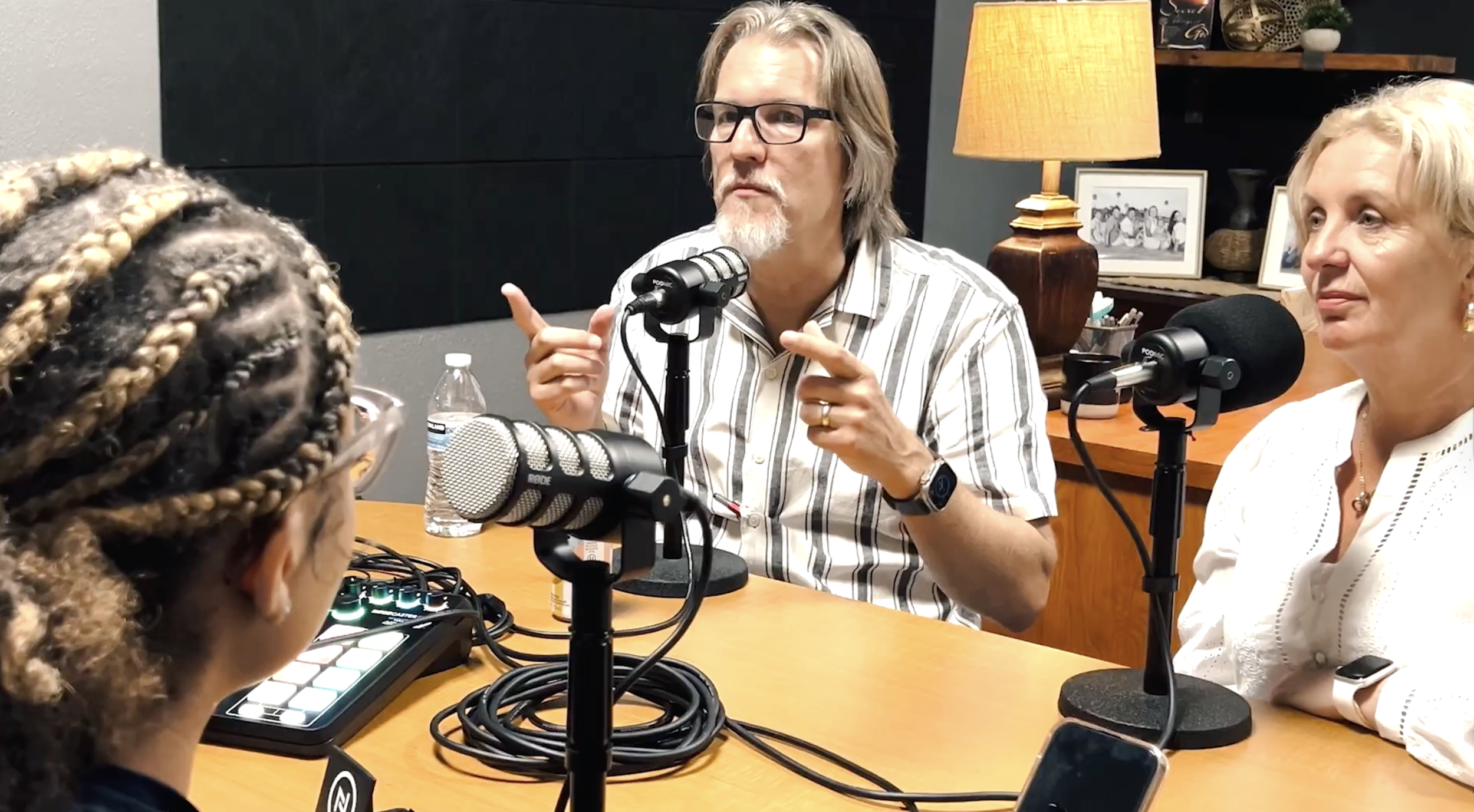 Three radio hosts sitting at a table with microphones, a sound mixer, and a water bottle, engaged in a discussion. One man with glasses and a beard points with his fingers, while a woman with blonde hair and a white blouse listens.