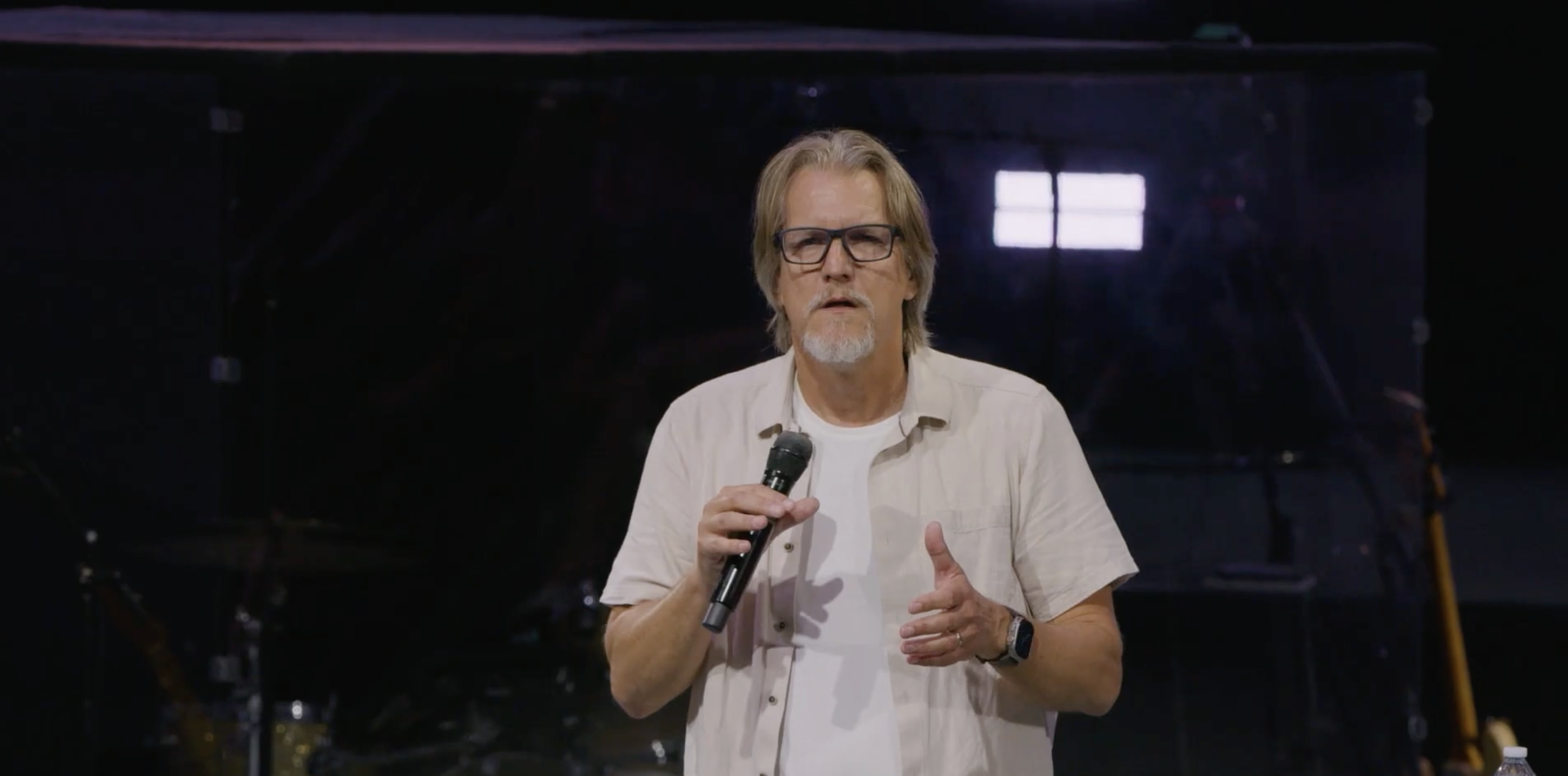 A man with glasses and gray hair, dressed in a white shirt, holding a microphone while speaking on stage with a dark background and window light.