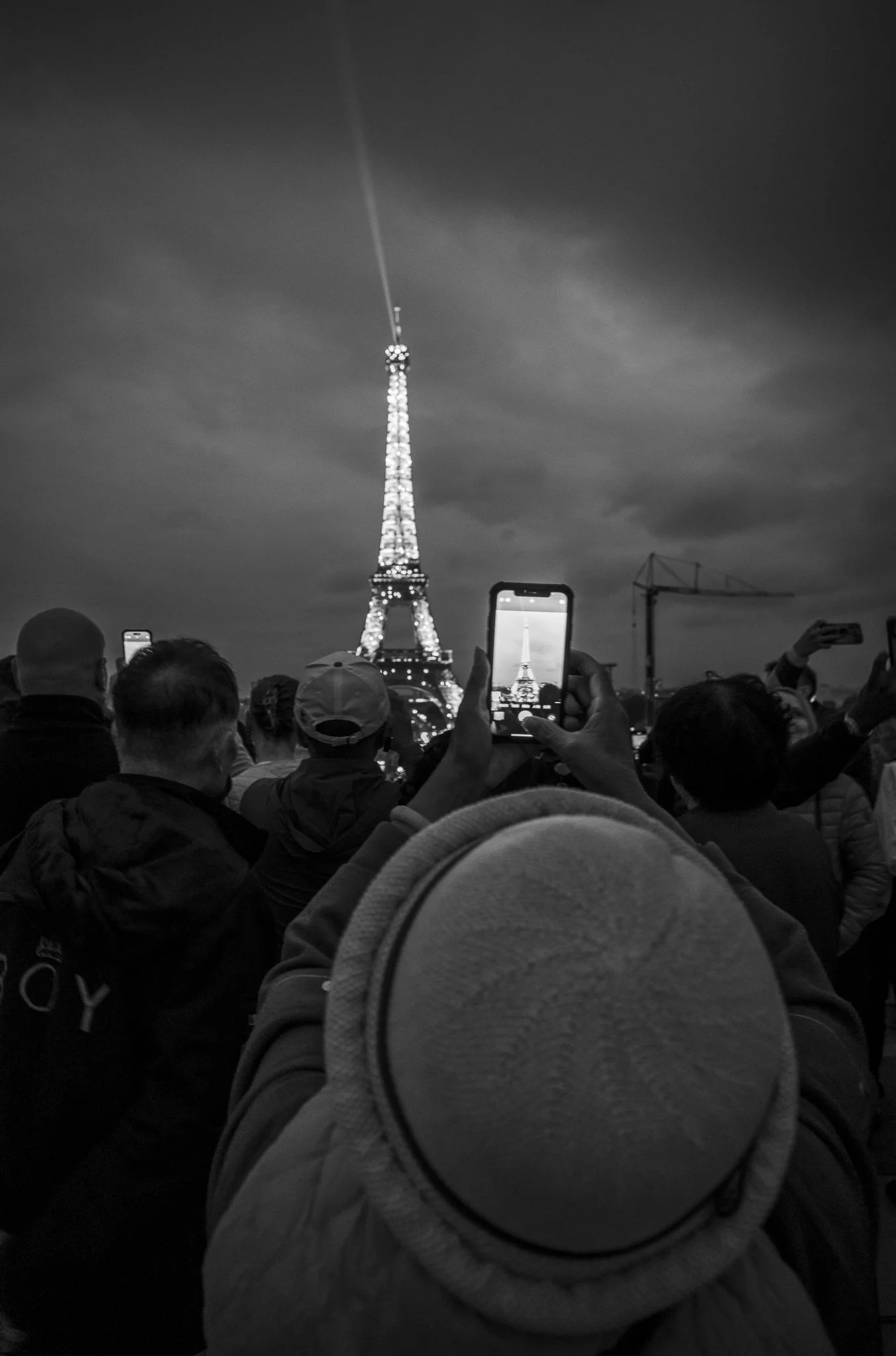 Menschen sammeln sich vor dem Eiffelturm in Paris, der nachts beleuchtet ist, Ein paar machen Fotos, während der Himmel bewölkt ist.