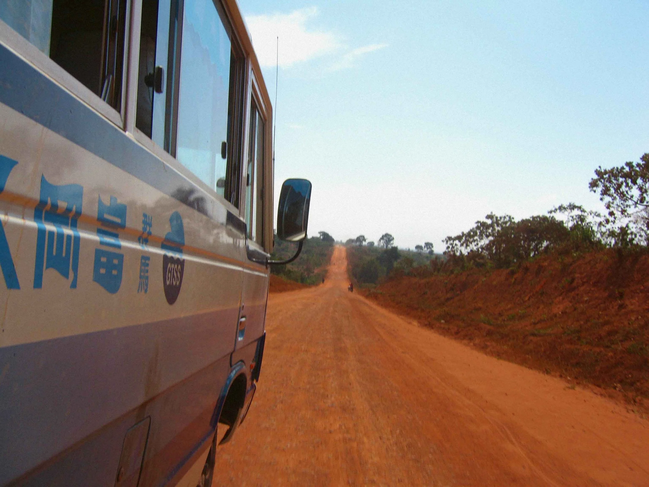 Part of a bus with Chinese characters on it, traveling on a dirt road through a rural landscape with trees and a clear sky.