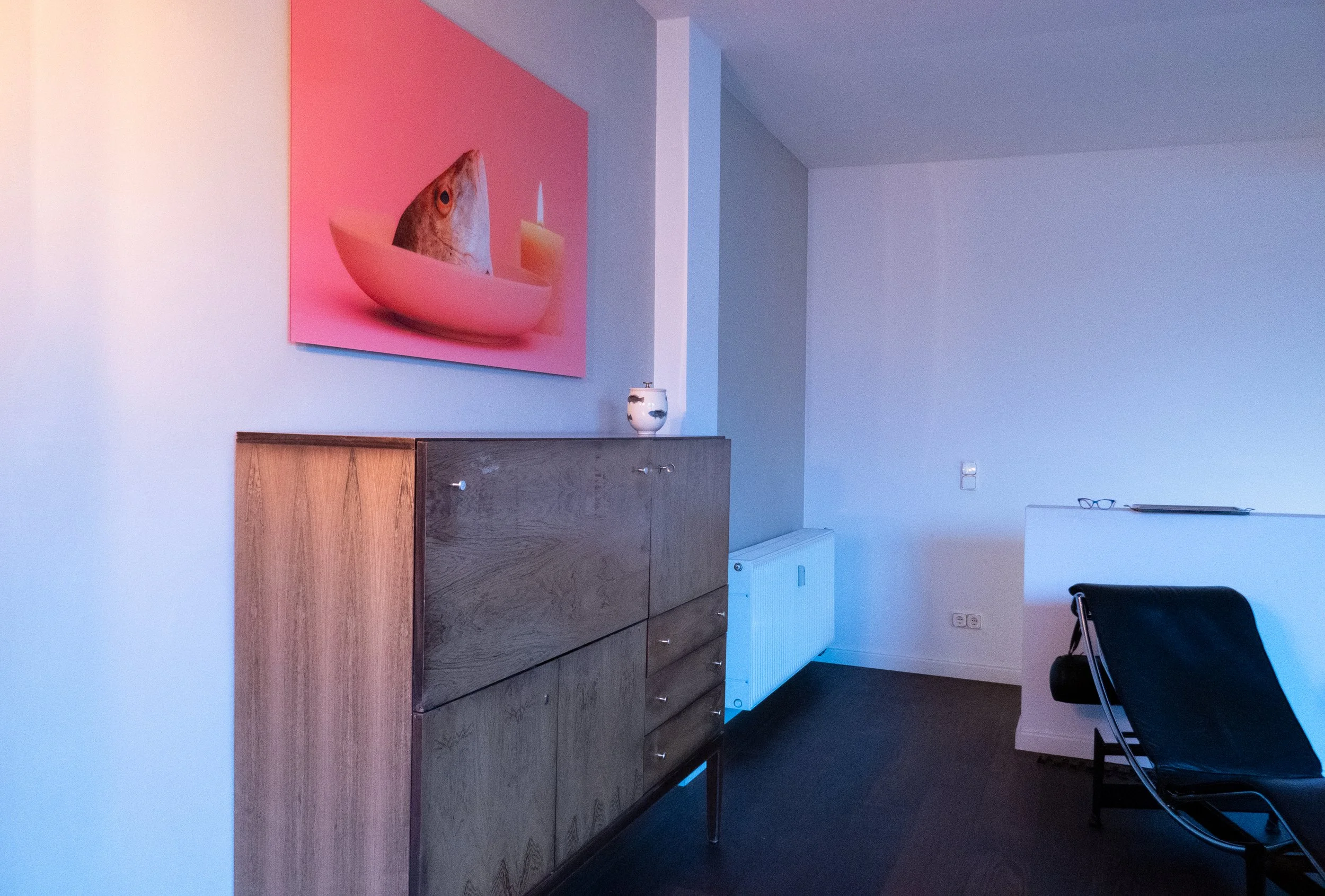 A room with a pink wall art featuring a fish head in a bowl with a candle, a wooden cabinet, a white radiator, a black chair, and a white desk with glasses and a laptop.