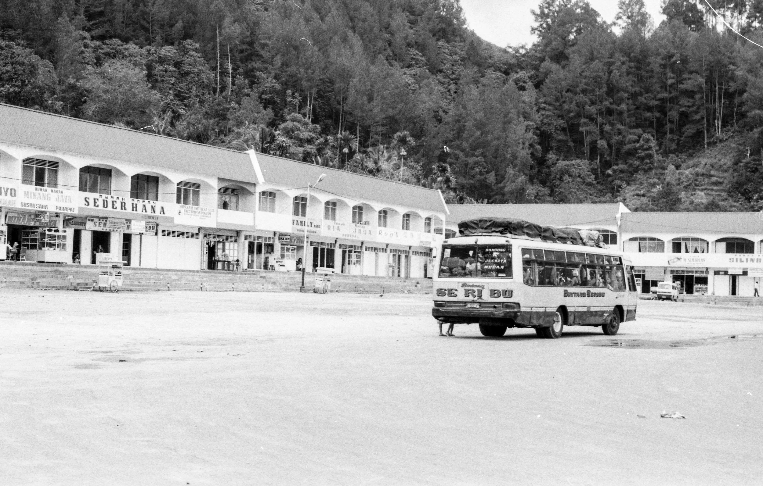 A black and white photo of a small bus standing alone in an open square with a mountainous backdrop. The bus has luggage on the roof and parked people inside.