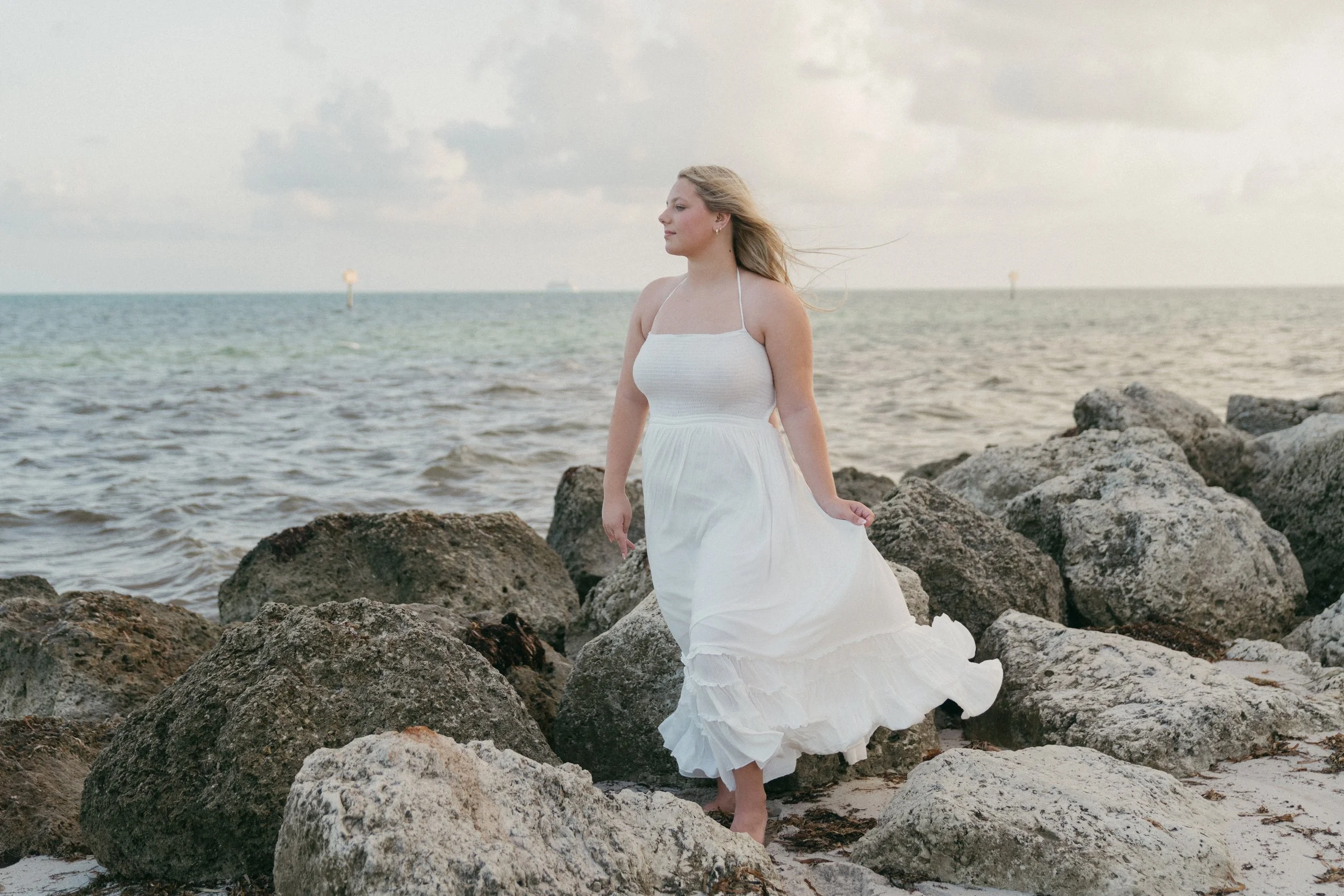 Woman in a flowing white dress posing on coastal rocks by the ocean during an outdoor portrait photography session in Key West, Florida.