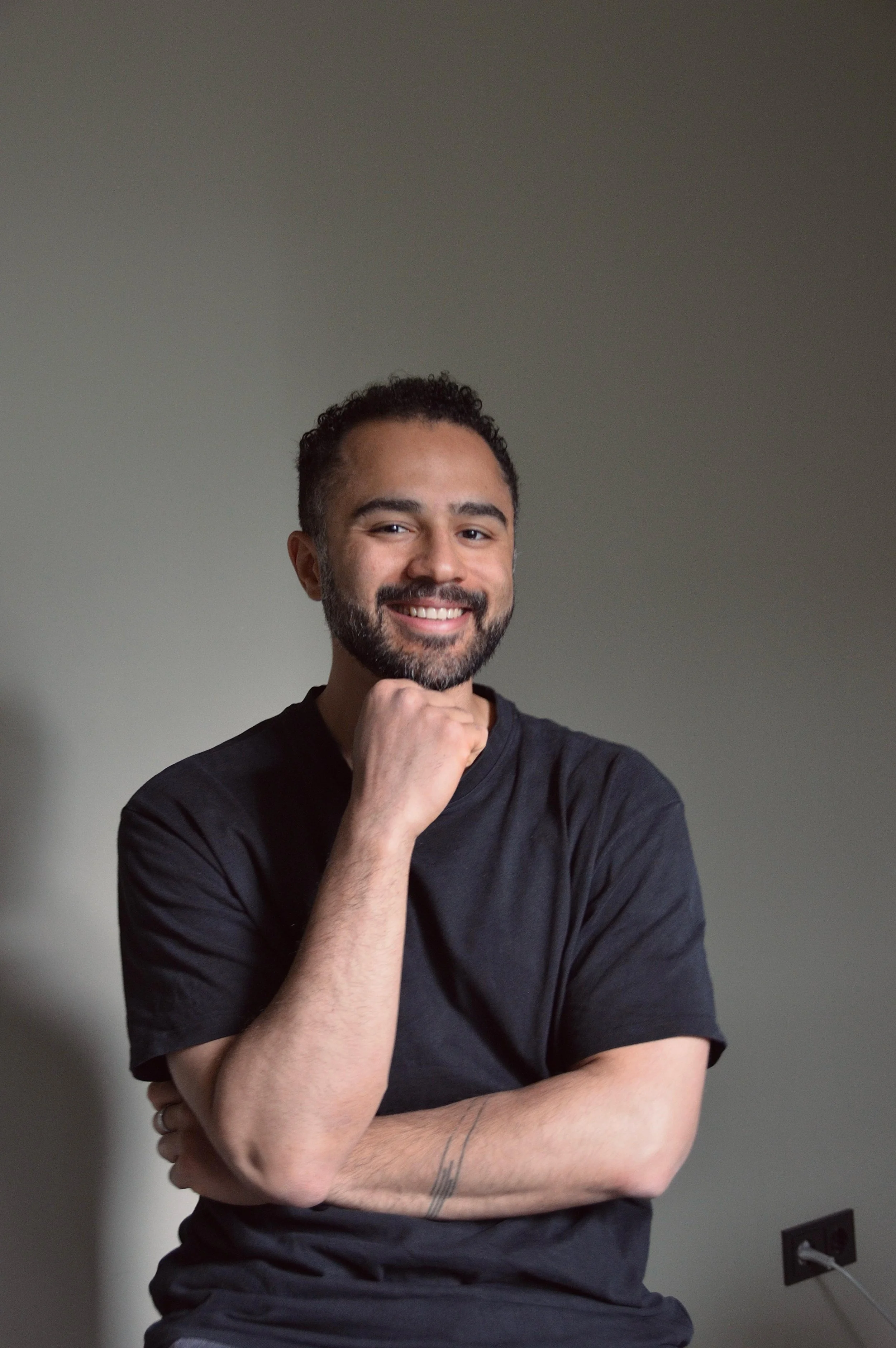Ein lachender Mann mit dunklem Bart und kurzen Locken, trägt ein schwarzes T-Shirt, sitzt vor einfachem grauem Hintergrund, mit einem Arm überkreuzt und der andere Hand, die seinen Kinn unterstützt.
