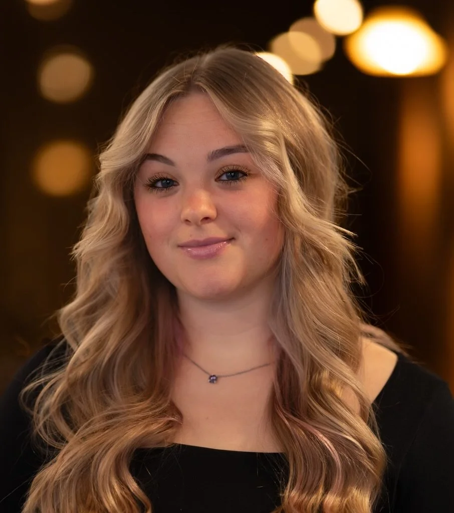 A young woman with wavy blonde hair, wearing a black top and a small necklace, smiling slightly against a blurred background with warm lights.