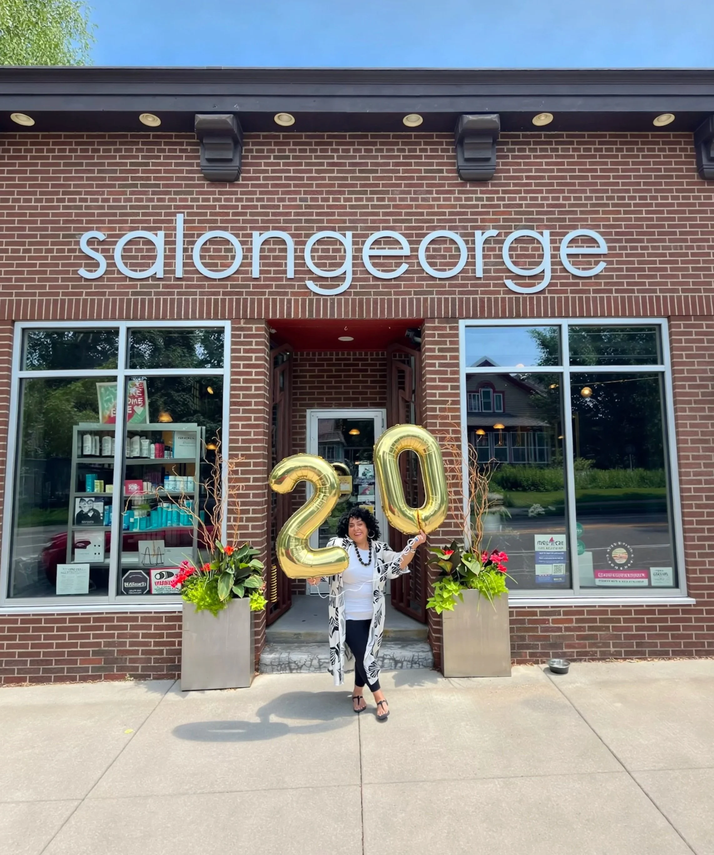 A woman with curly black hair is standing in front of a brick storefront holding large gold number balloons ’2’ and ’0’ for a 20th celebration. She is dressed in a white outfit with a black and white animal print scarf and black high heels. The store behind her has large windows with displays and the word ‘salongeorge’ on the brick wall above the entrance. There are potted plants with flowers on either side of the entrance, and a large gold ‘20’ balloon arrangement is visible.