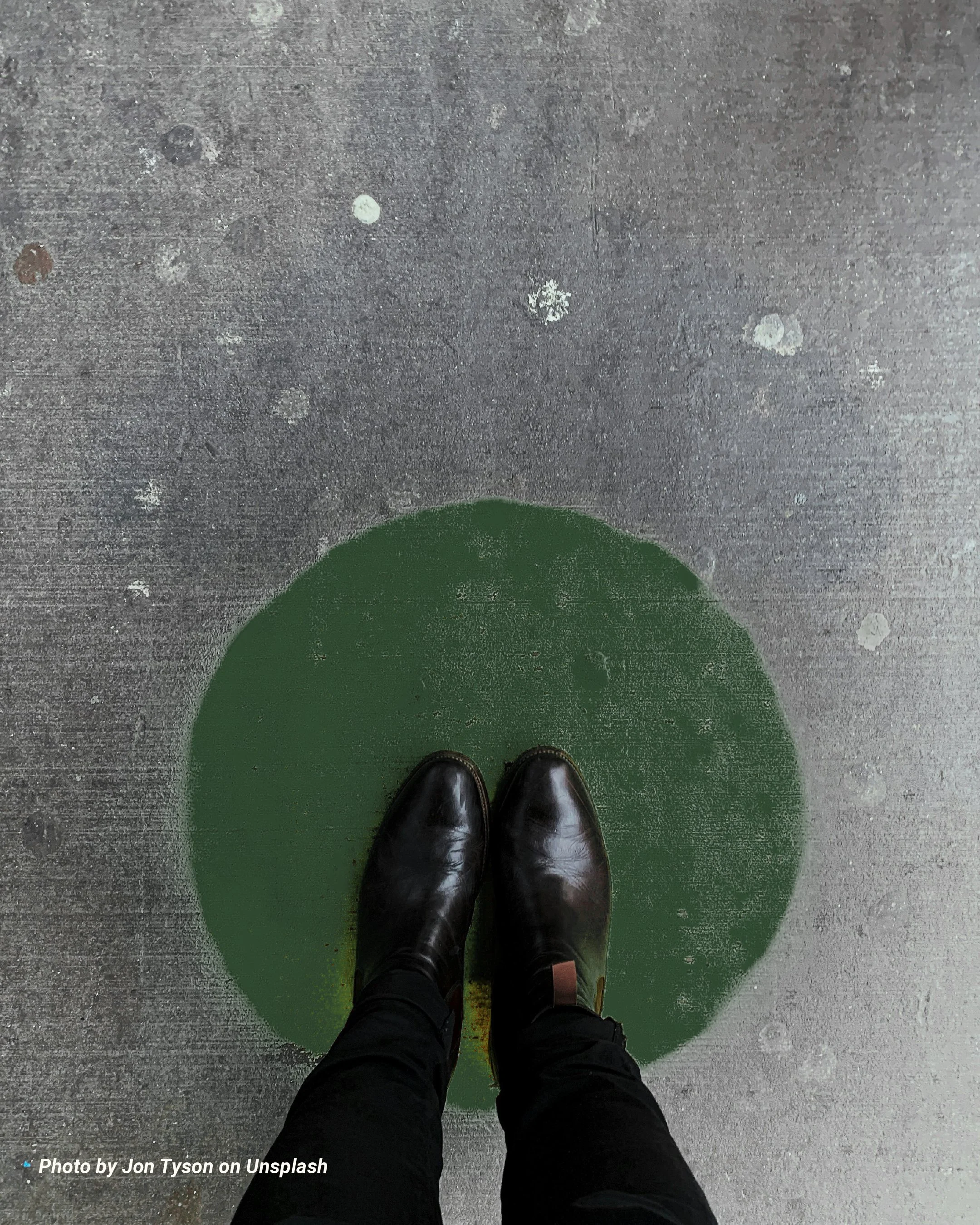 A person wearing black shoes and black pants standing on a gray concrete floor with a green painted circle underneath.