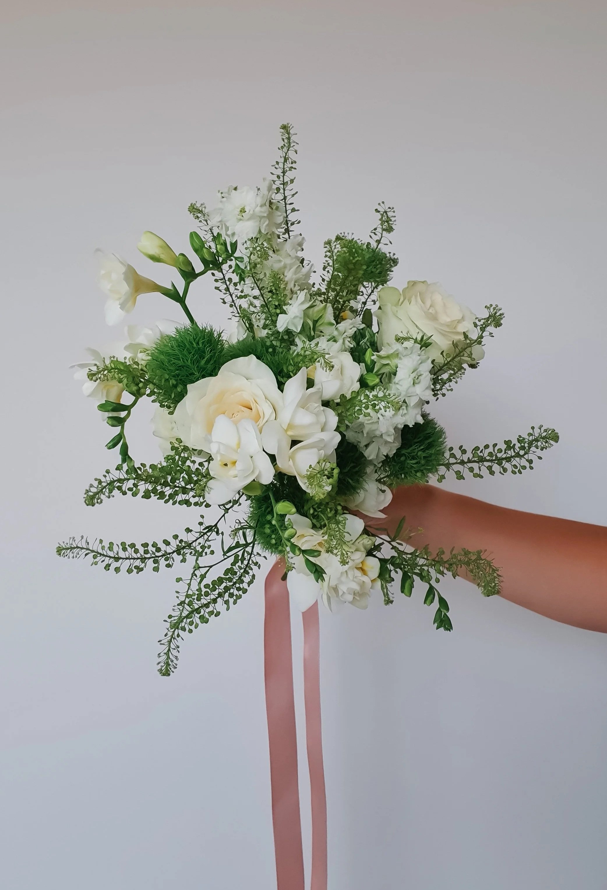 Een hand die een wit bruidsboeket vasthoudt met lange roze linten tegen een effen lichte achtergrond.