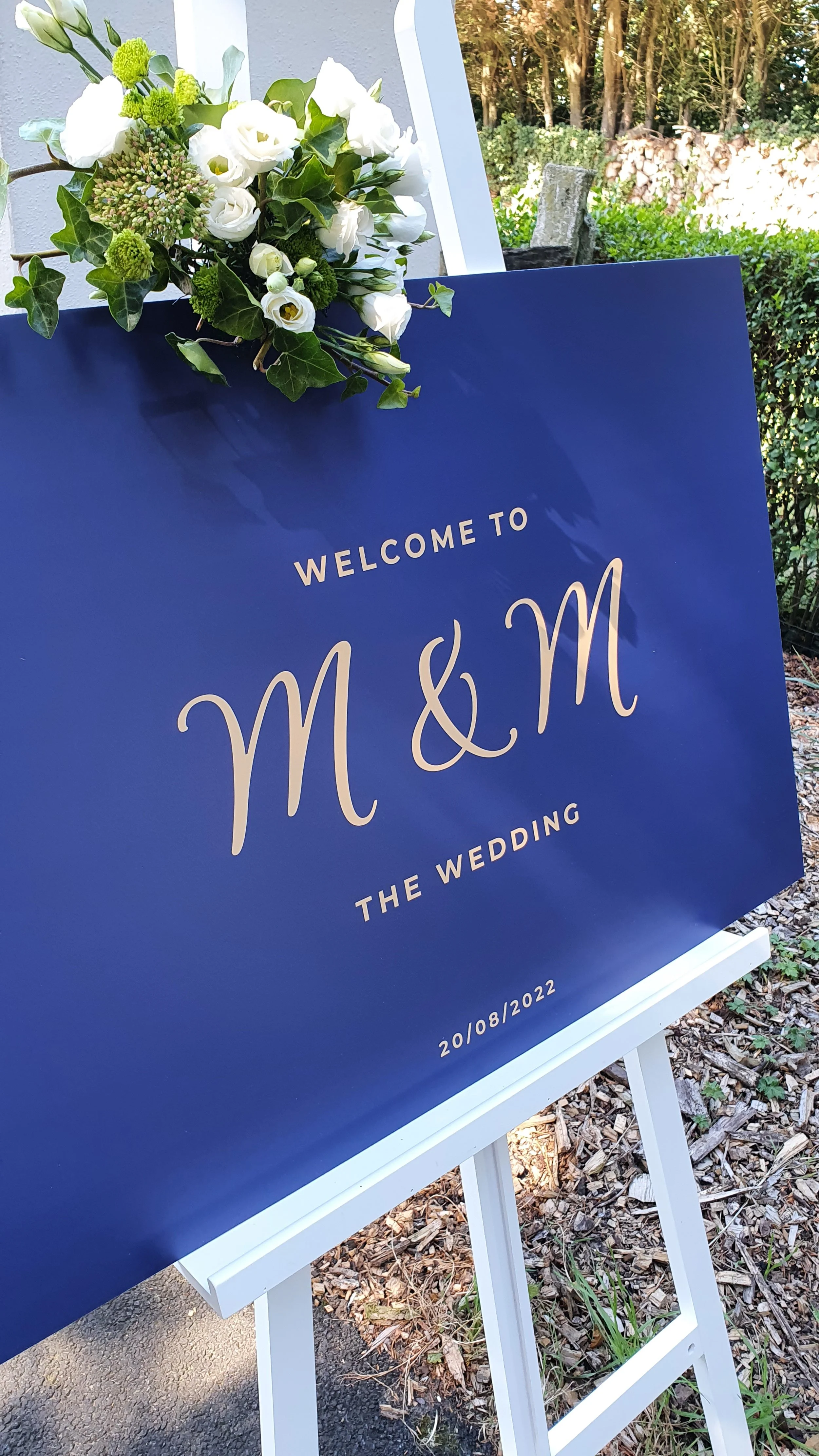 Bord met tekst: 'Welkom bij M&M het huwelijk' en datum 20/08/2022, versierd met een bloemarrangement bestaande uit witte bloemen en groene bladeren, buiten opgesteld op een witte standaard.