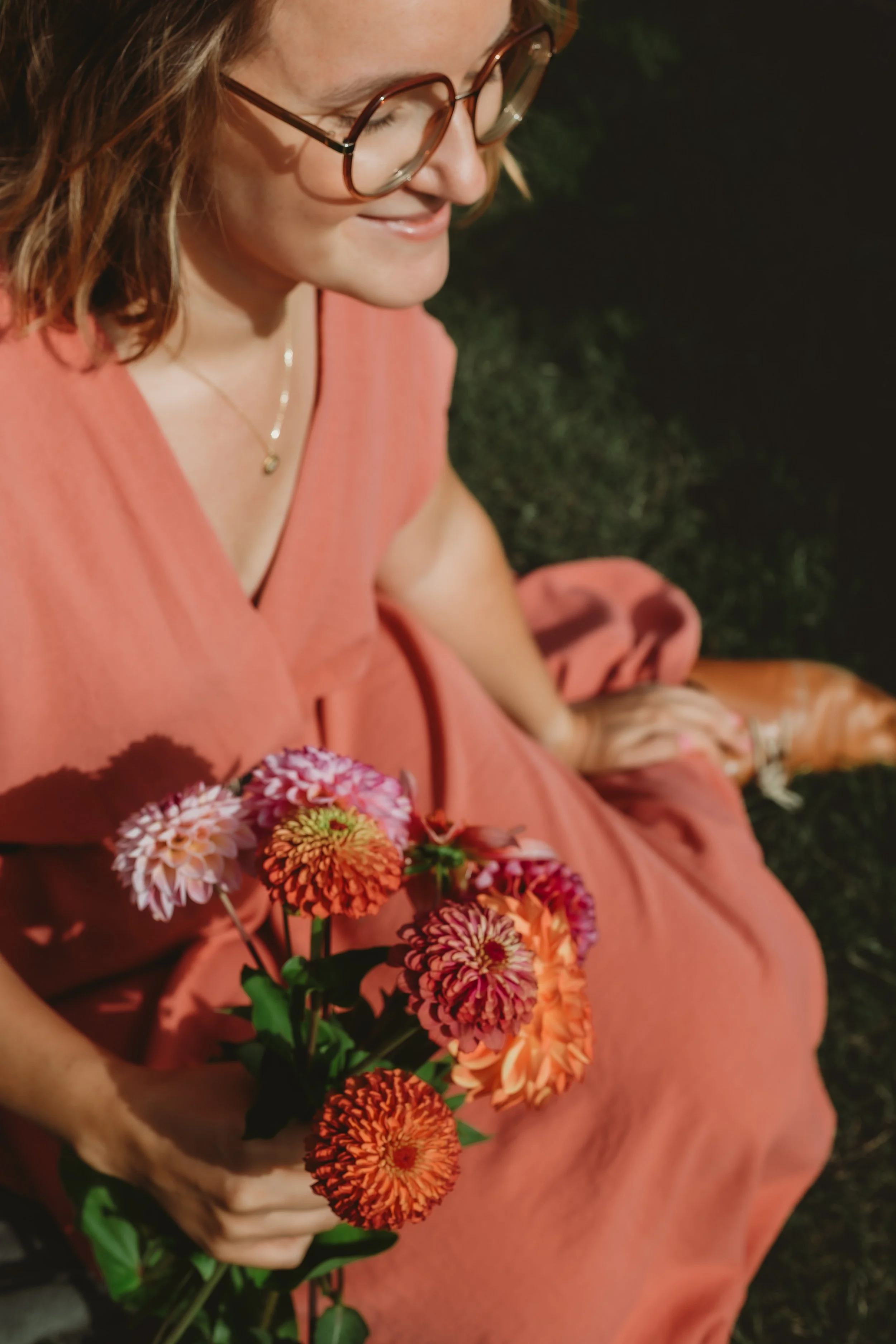 Close-up van een lachende vrouw met bruin haar en bril, zittend op gras, met een boek en een bos bloemen in haar handen, gekleed in een roze jurk.