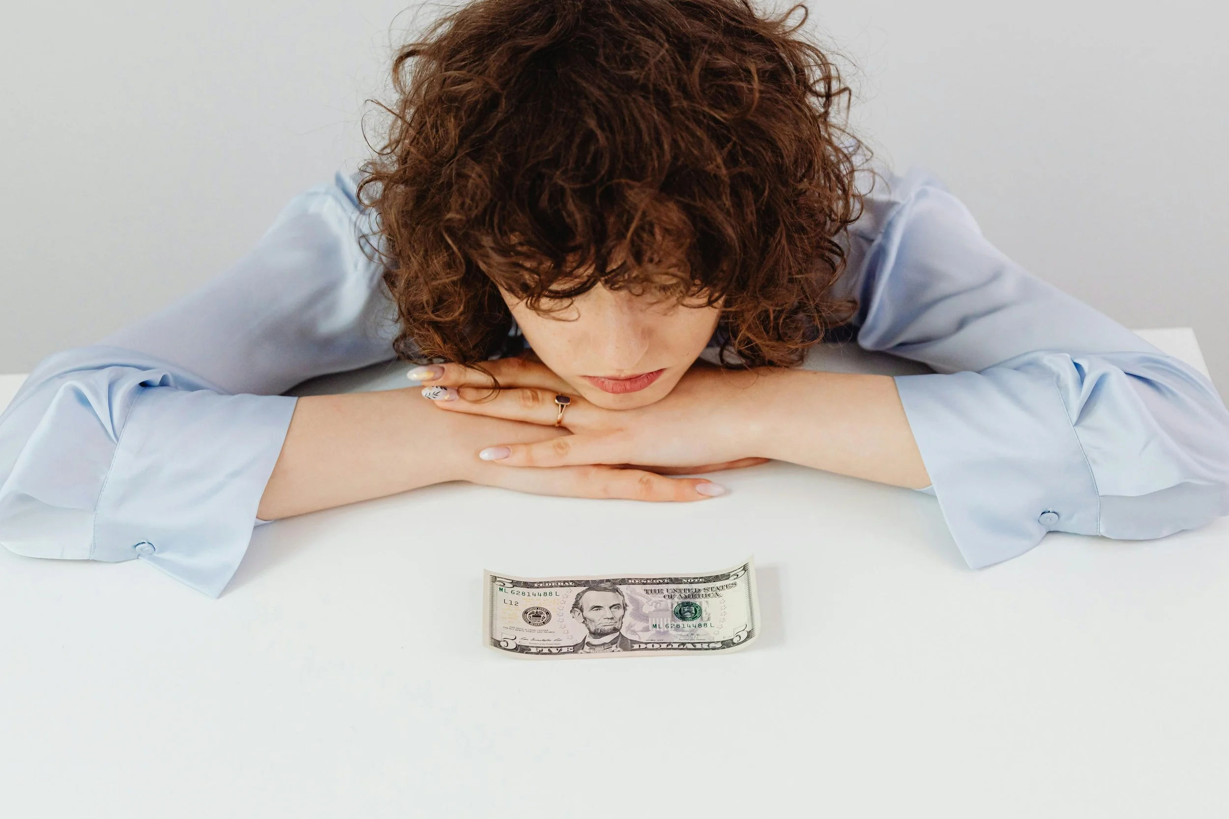 Woman with curly brown hair wearing a light blue button-up shirt rests her head on her folded arms on a white surface, looking down at a five-dollar bill placed in front of her with a contemplative or concerned expression.
