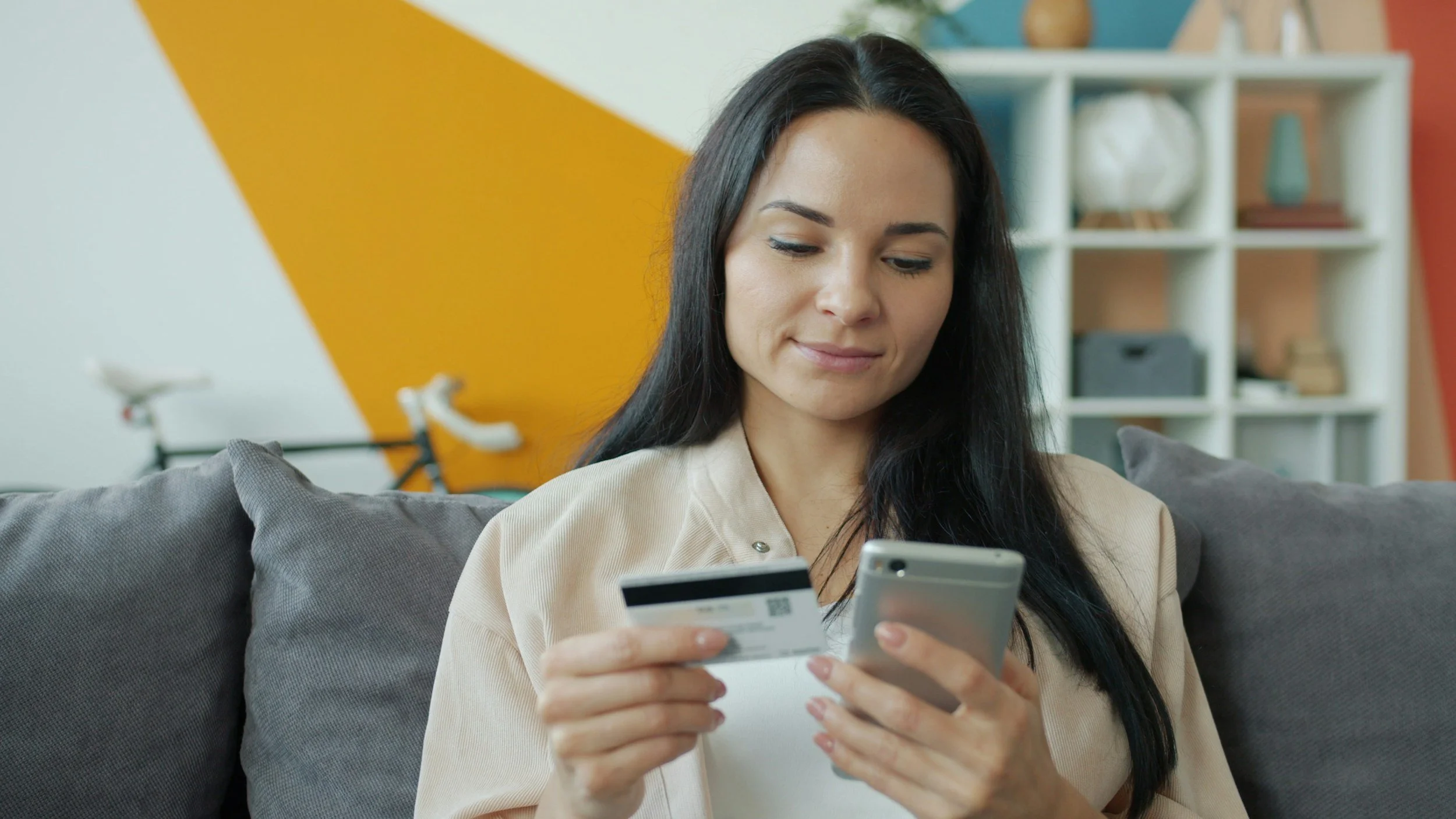 Young woman with long dark hair wearing a cream-colored top sits on a gray couch, smiling while holding a credit card in one hand and looking at her phone in the other hand, with a bright modern living room and white shelving in the background.