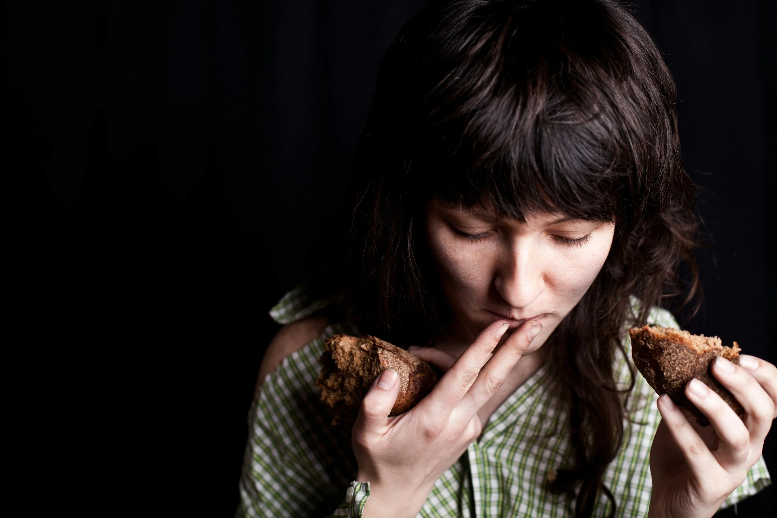 A woman with dark hair eating a piece of bread with both hands, eyes downcast, against a dark background.