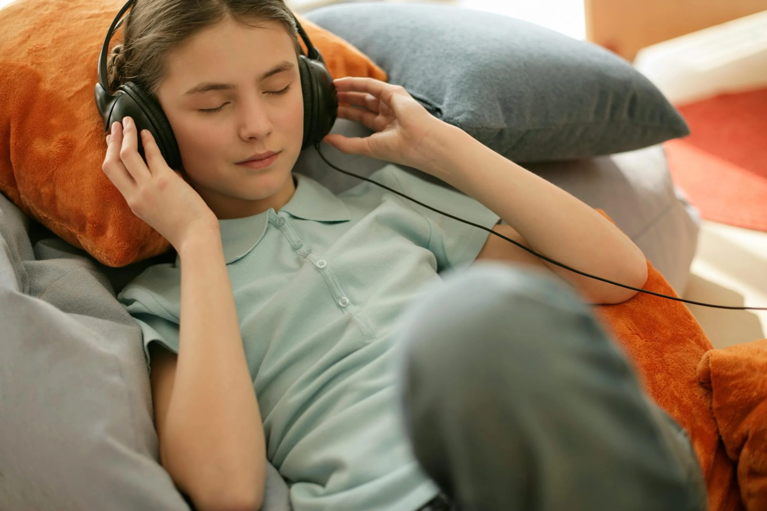 A young person lying back on colorful pillows with eyes closed, wearing over-ear headphones and resting one hand gently on the side of their head, appearing calm and relaxed.