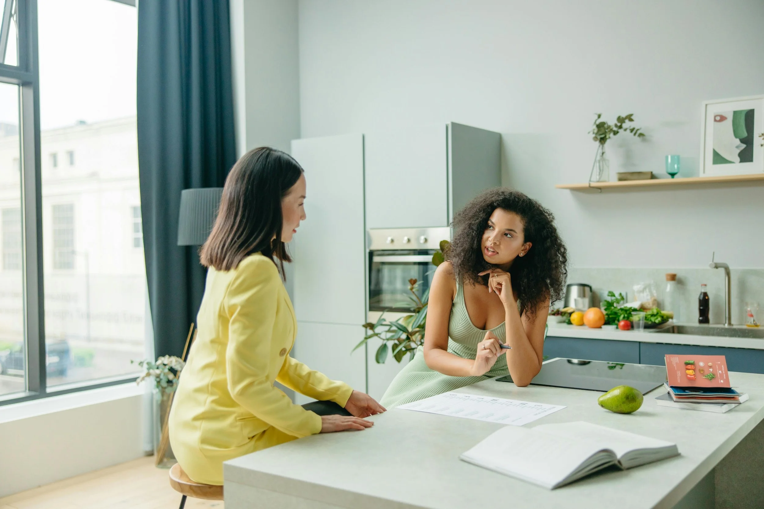 Two women having a conversation at a kitchen counter, one in a yellow blazer and one in a sage green top, with a notebook, fruit, and natural light visible in the background.
