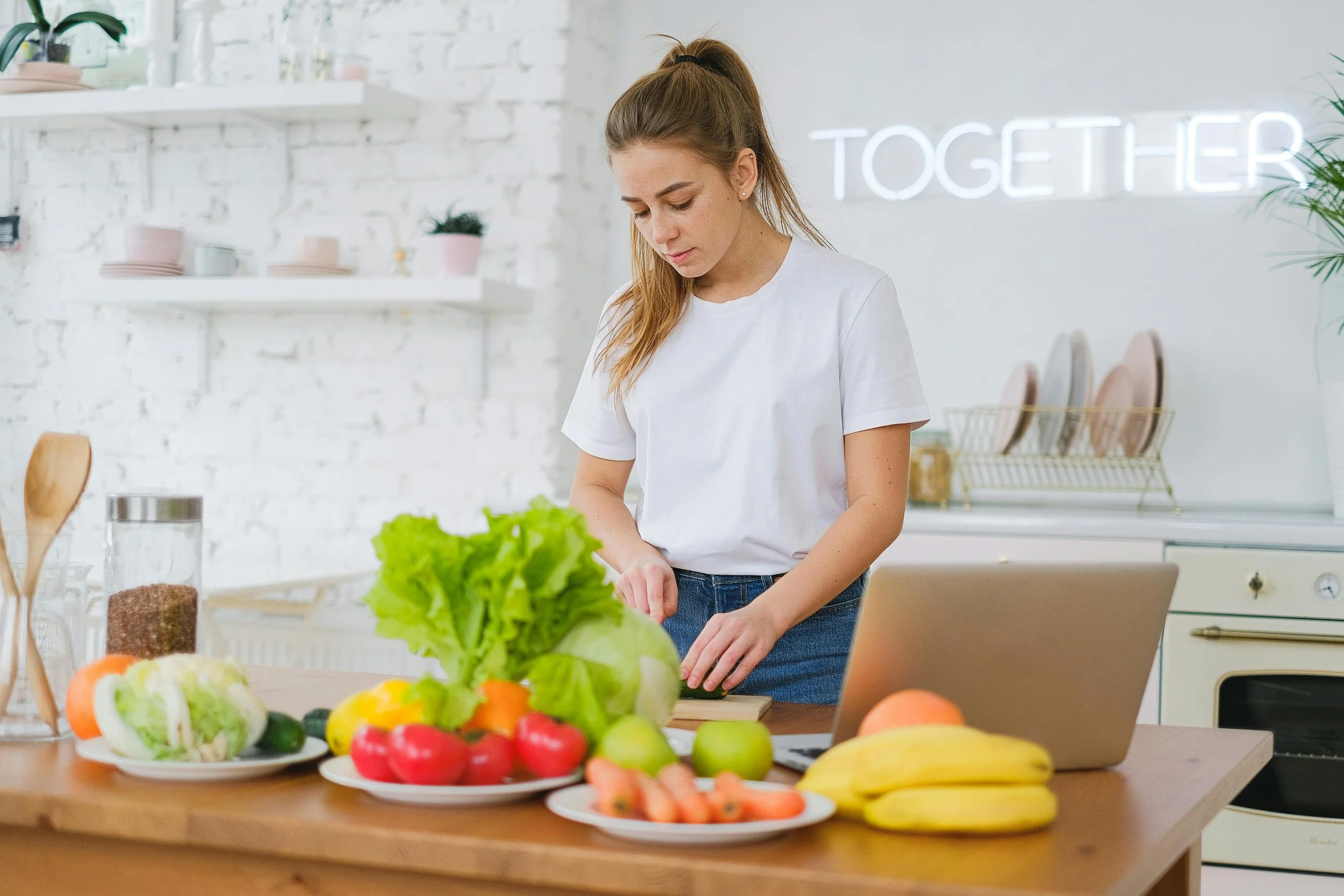 A young woman in a white t-shirt stands at a kitchen counter with a laptop, surrounded by fresh vegetables and fruit including lettuce, bell peppers, carrots, apples, and bananas. A neon sign reading 'TOGETHER' glows on the wall behind her.