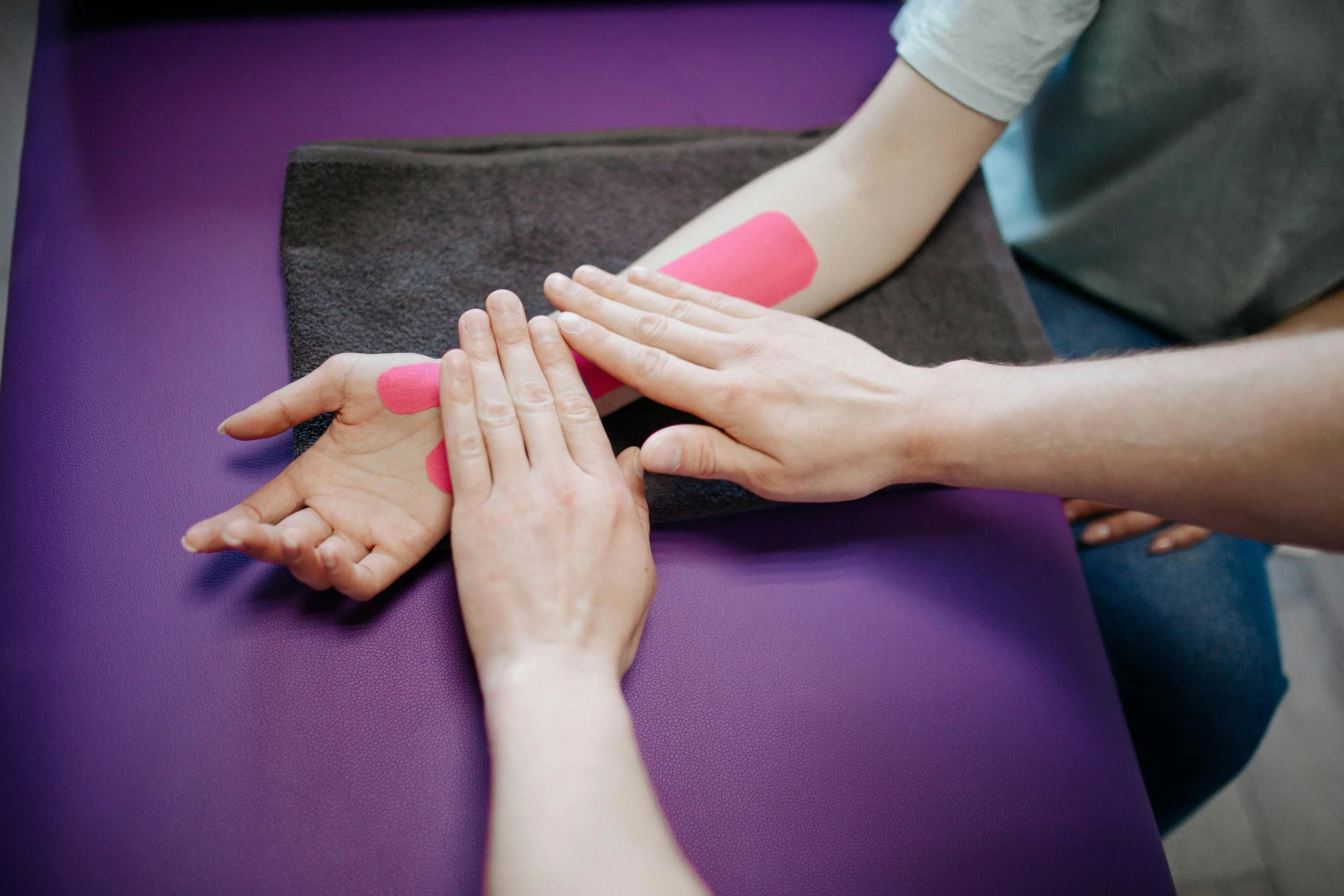 A physical therapist's hands gently guide a patient's hand and wrist on a purple mat, with pink kinesiology tape visible on the patient's forearm.
