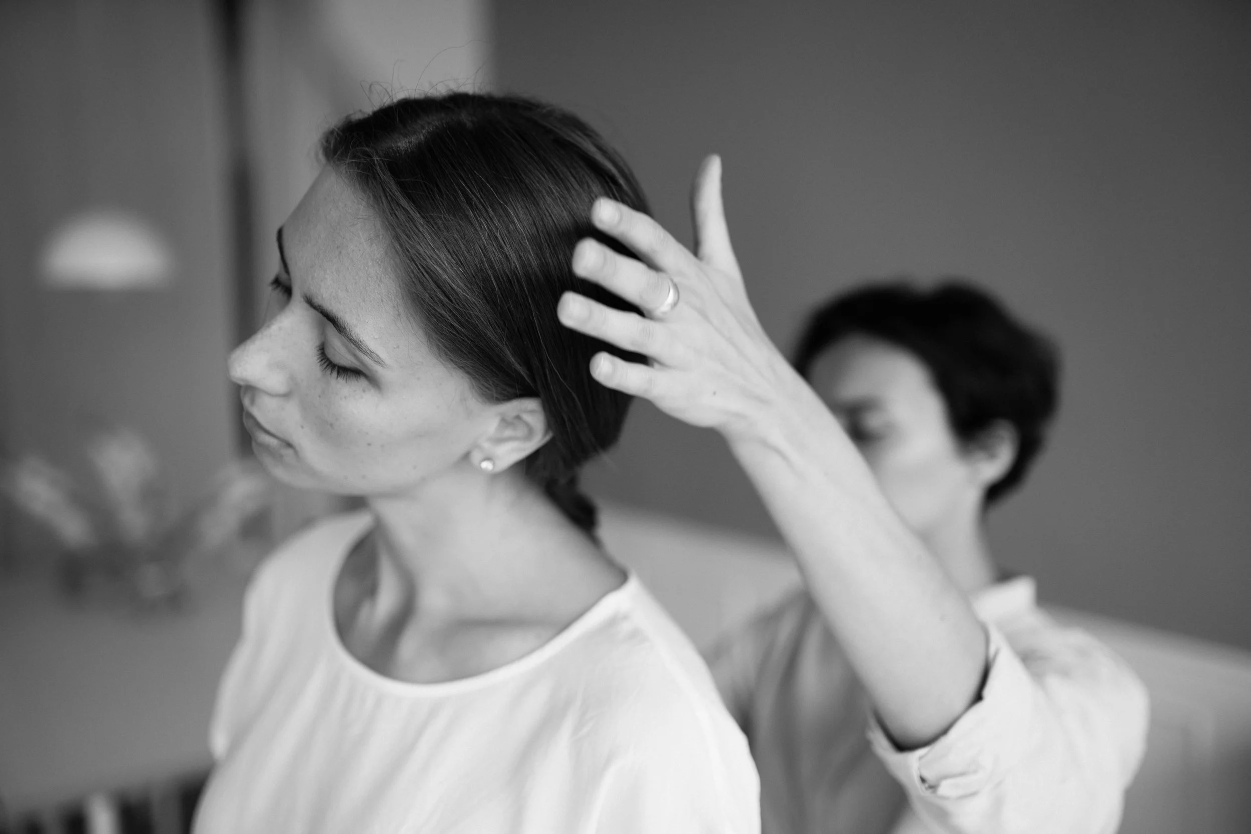 A black and white photo of two women in a therapy session. A woman with dark hair pulled back stands in the foreground with her eyes closed, while the other woman stands behind her supporting her head leaning slightly to the left.