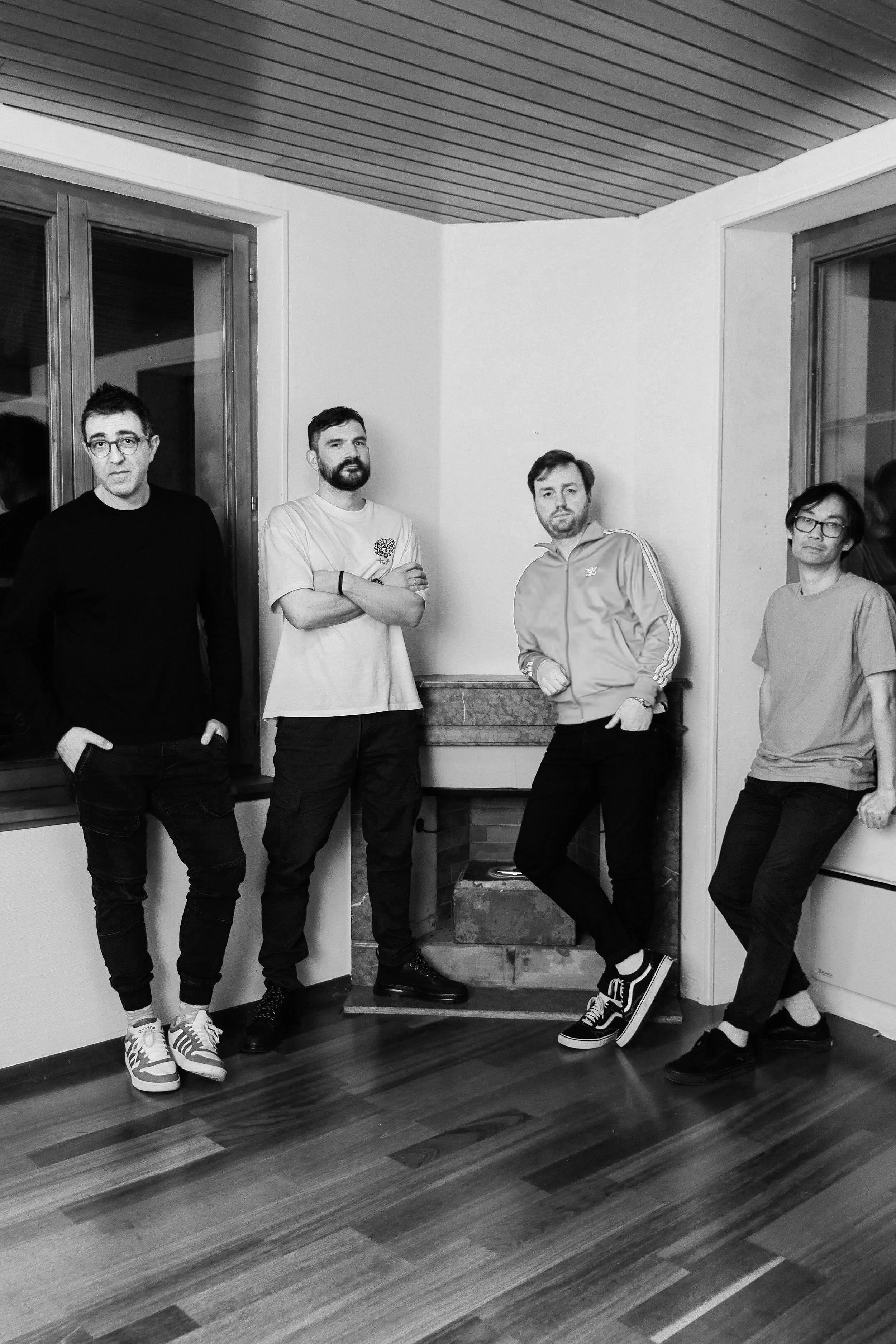 Four men standing in a room with wooden floors and walls, facing the camera with serious expressions. They are dressed casually, some with arms crossed or hands in pockets.