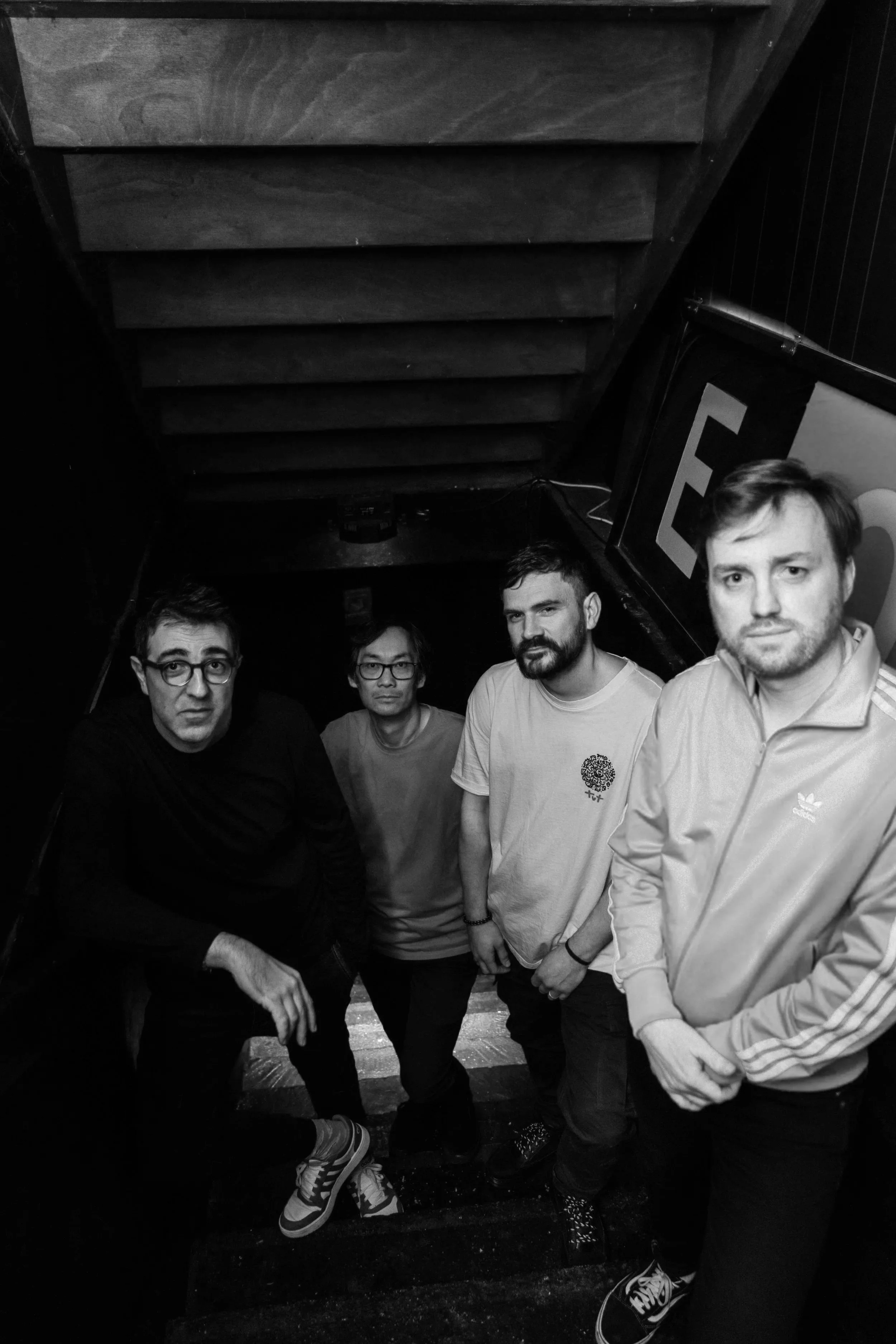 Five men standing on a staircase in a dark indoor setting, with one person partially out of the frame, looking at the camera.