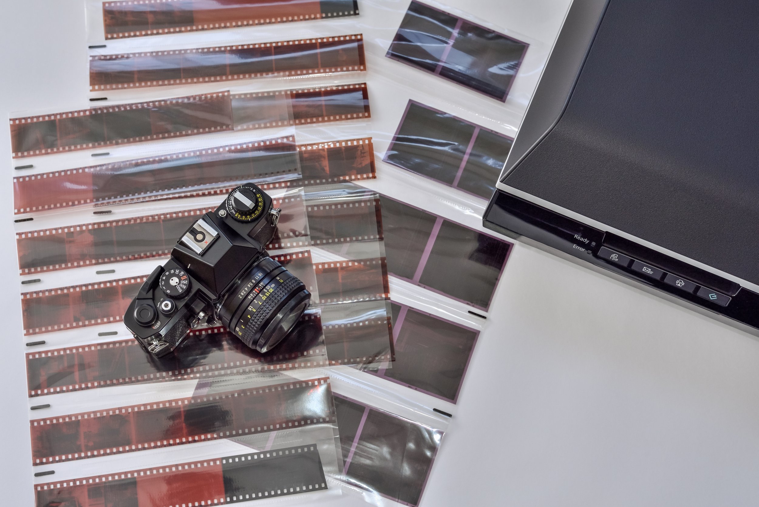 A film camera, negative strips, and photo prints on a white surface, with part of a photographic printer or scanner visible.
