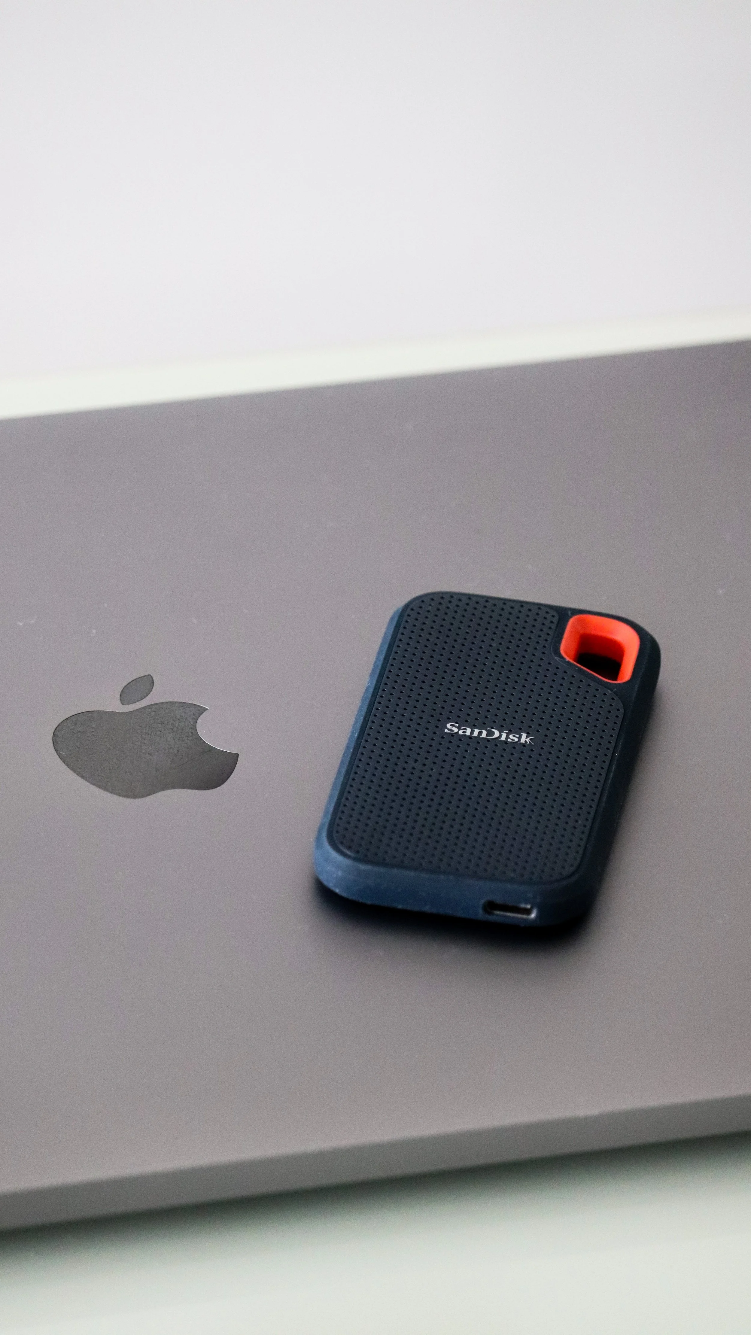 A black SanDisk portable external hard drive resting on the lid of a closed silver MacBook laptop, with the Apple logo visible, on a white surface.