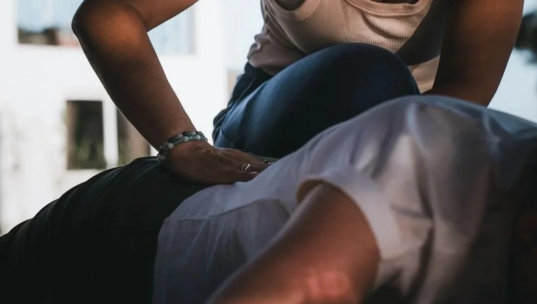 Yoga Assist indem eine Hand auf dem unteren Rücken einer Schülerin gelegt wird um dort zu entspannen