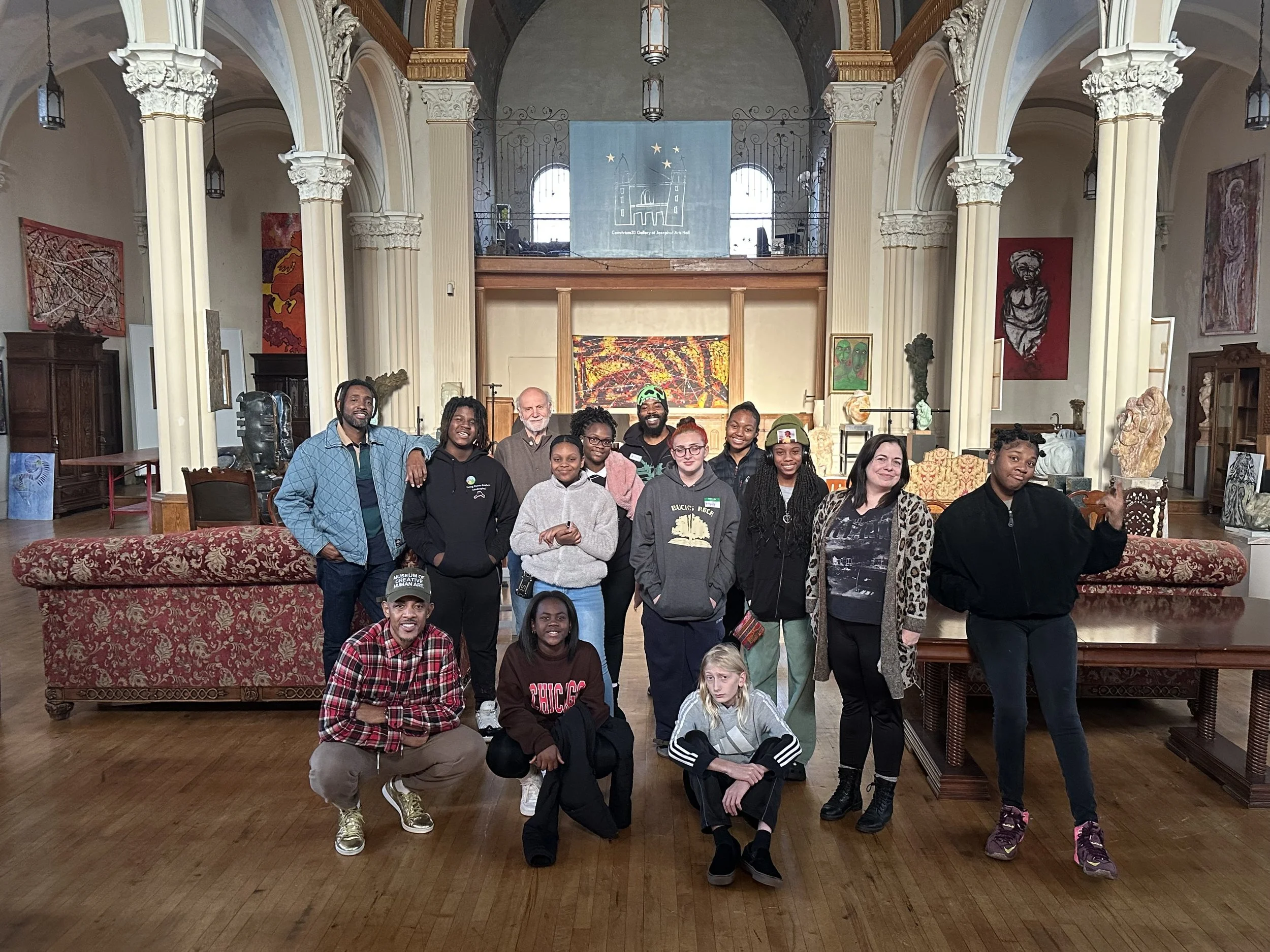 Group of diverse people posing inside an art gallery or historic building with high ceilings, arched windows, and various artwork displayed on the walls.