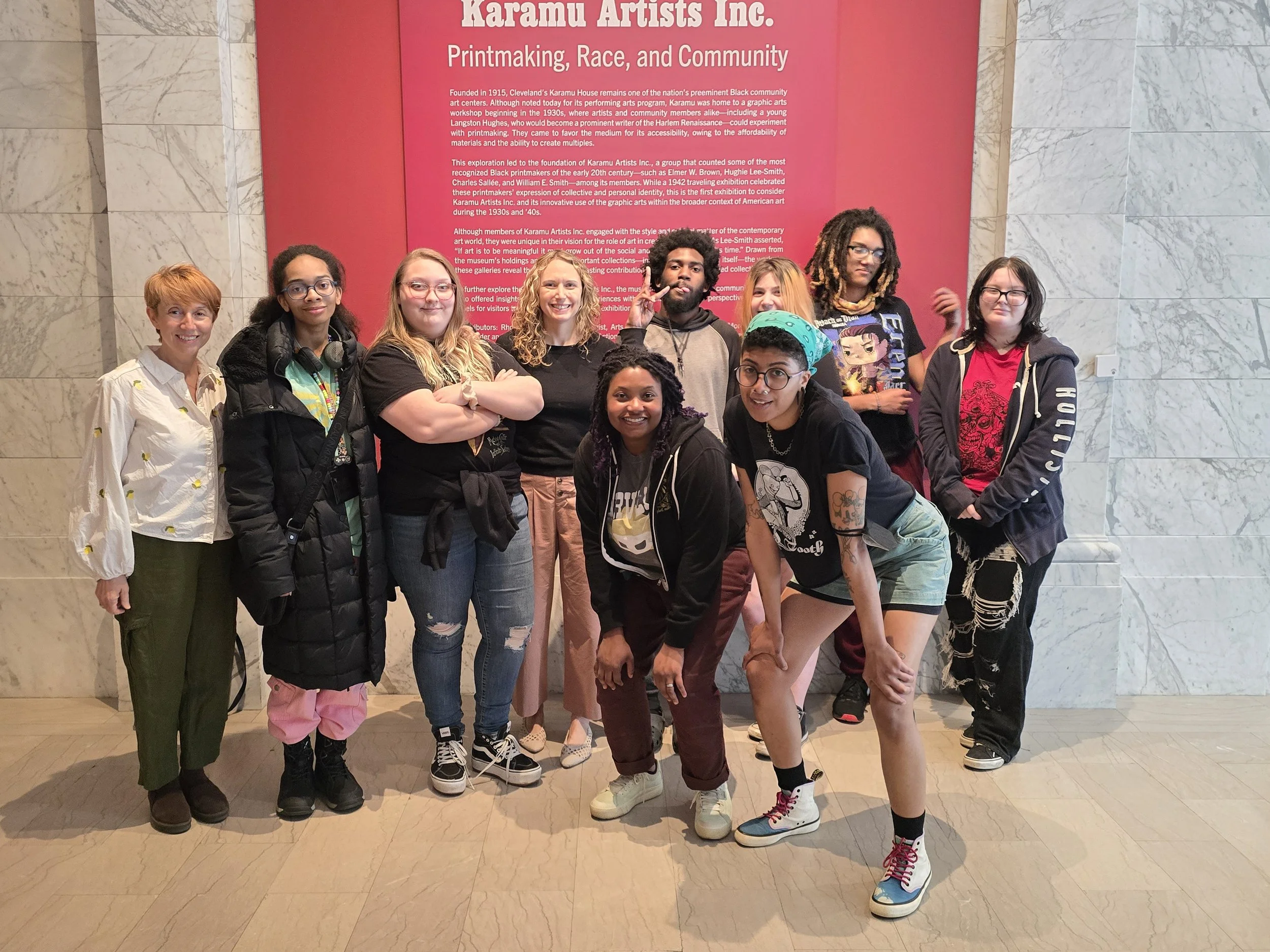 Group of nine people standing in front of a red informational wall, smiling and posing for a photo. The wall has text about Karamu Artists Inc. and historical information.