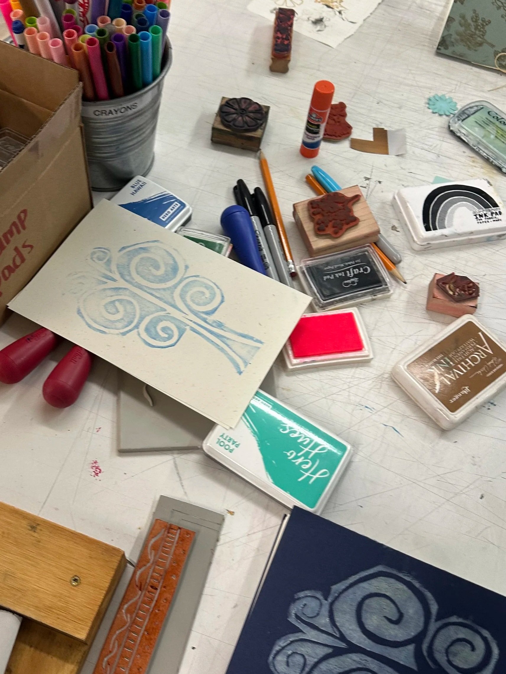 A cluttered workspace with various craft supplies, including a container of colorful markers, ink pads, stamps, glue stick, paper, and a wooden box, on a white table with a blue patterned rug in the background.