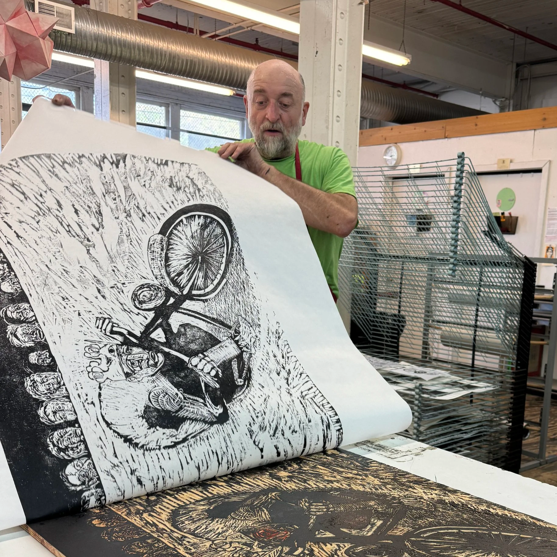 A man with a beard in a green shirt examining a large black and white print of a graphic artistic depiction of a skull with lemon slices, in an art studio or workshop with racks and industrial lighting.