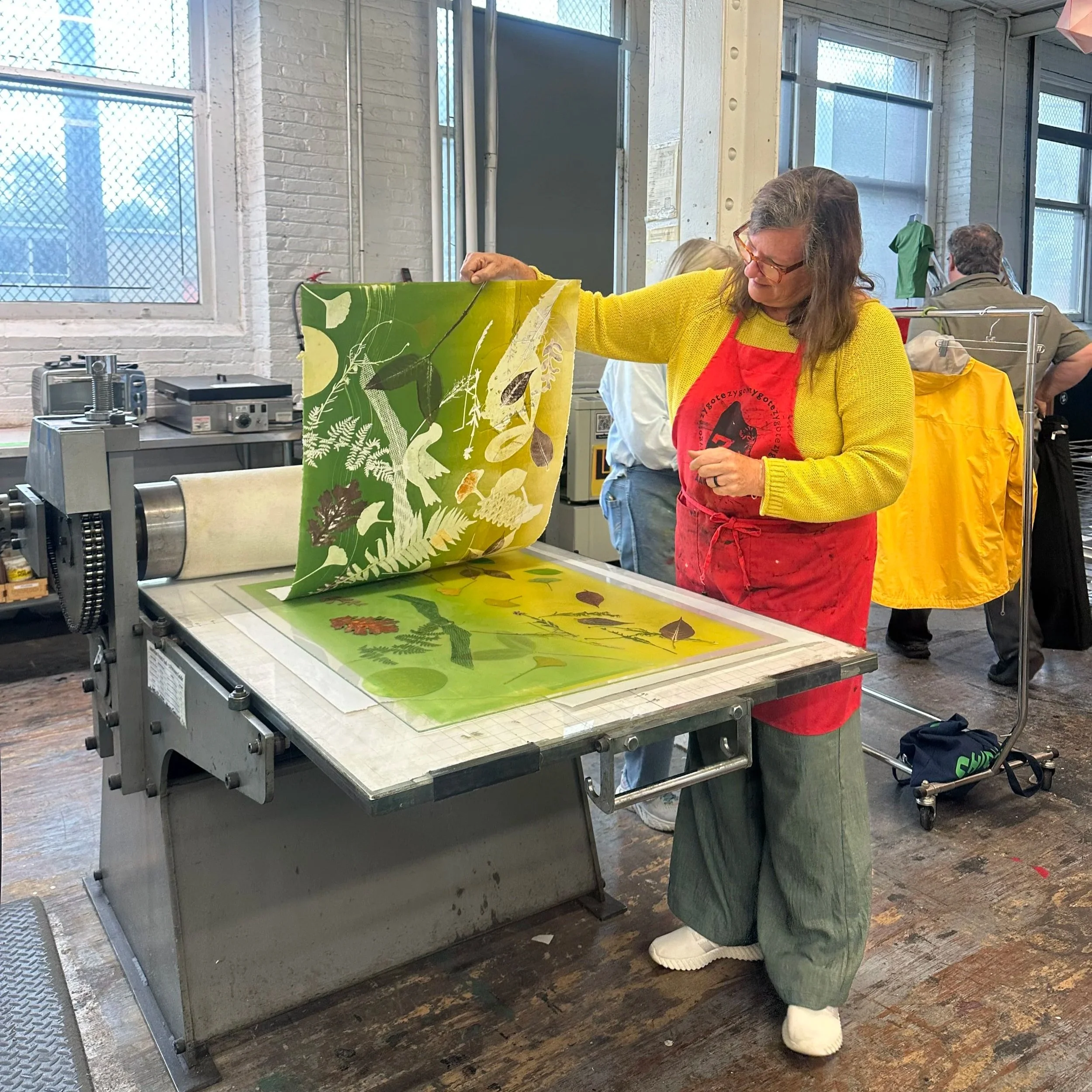 A woman with glasses, a yellow sweater, and a red apron is working with a large piece of green fabric with botanical designs in an industrial workspace. She is holding the fabric and appears to be preparing or inspecting it. In the background, a man with glasses, a grey shirt, and a yellow apron is looking to the side. The workspace has large windows with mesh screens and various equipment on the table.