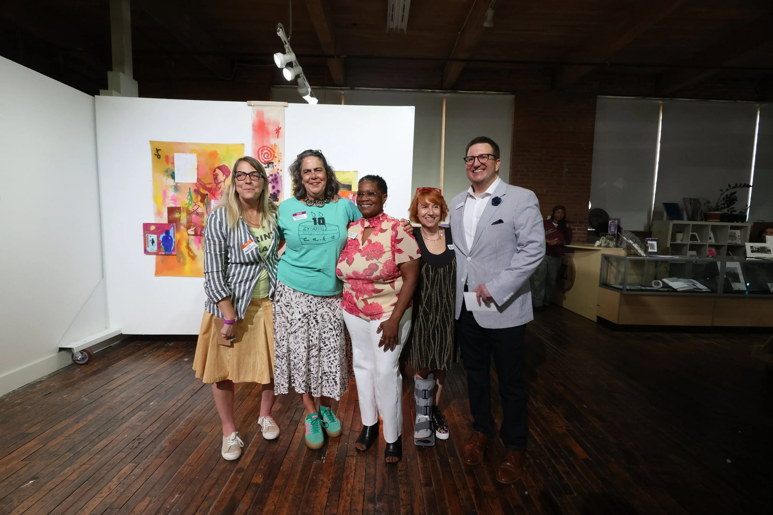 Five people standing together in an art gallery, smiling for the camera. They are dressed in casual and semi-formal attire. The background features colorful abstract artwork and a white wall.