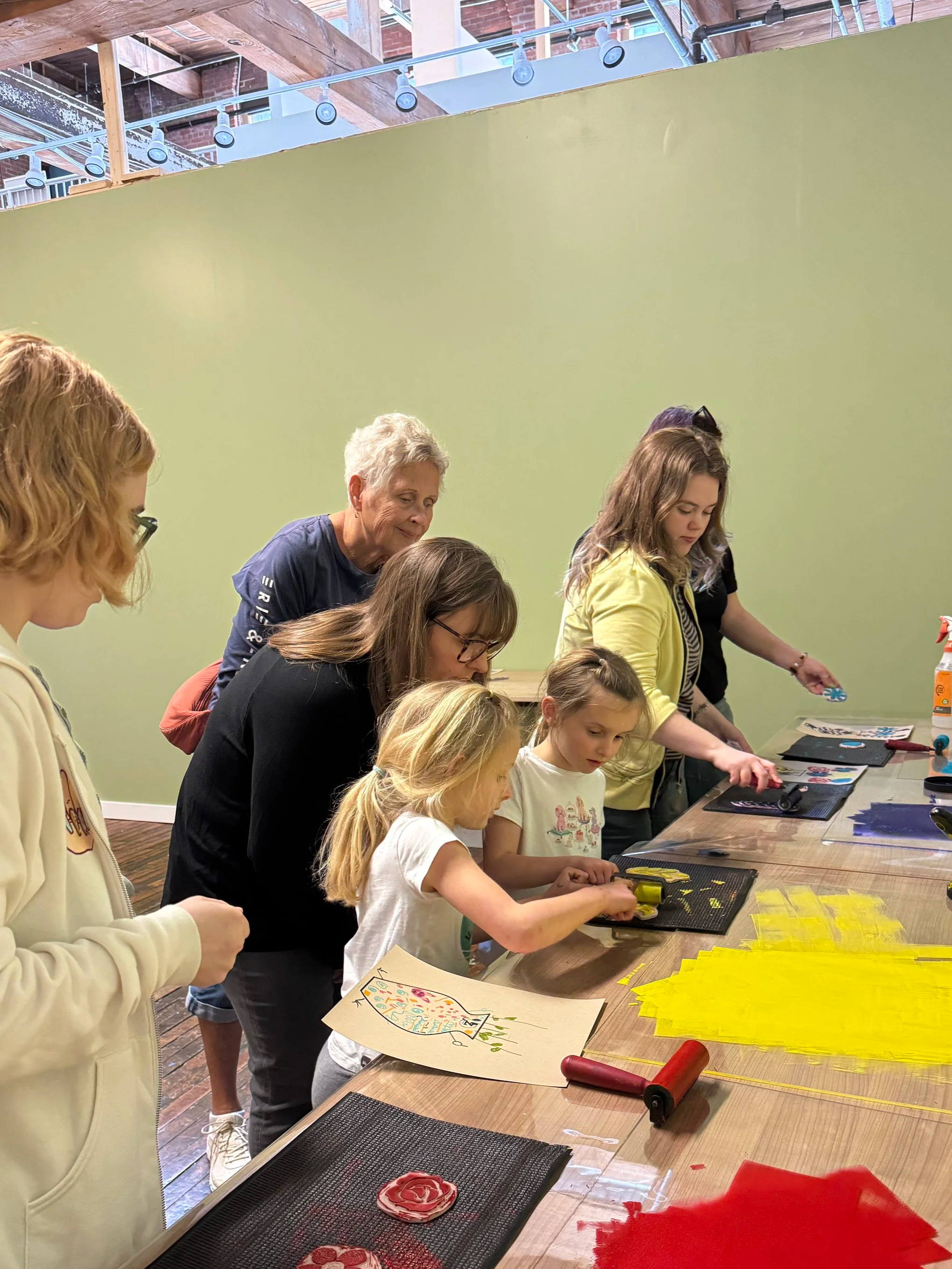 Group of children and adults participating in an arts and crafts activity at a long table, with colorful paper and craft supplies, in a room with a green wall and exposed brick and wooden beams ceiling.