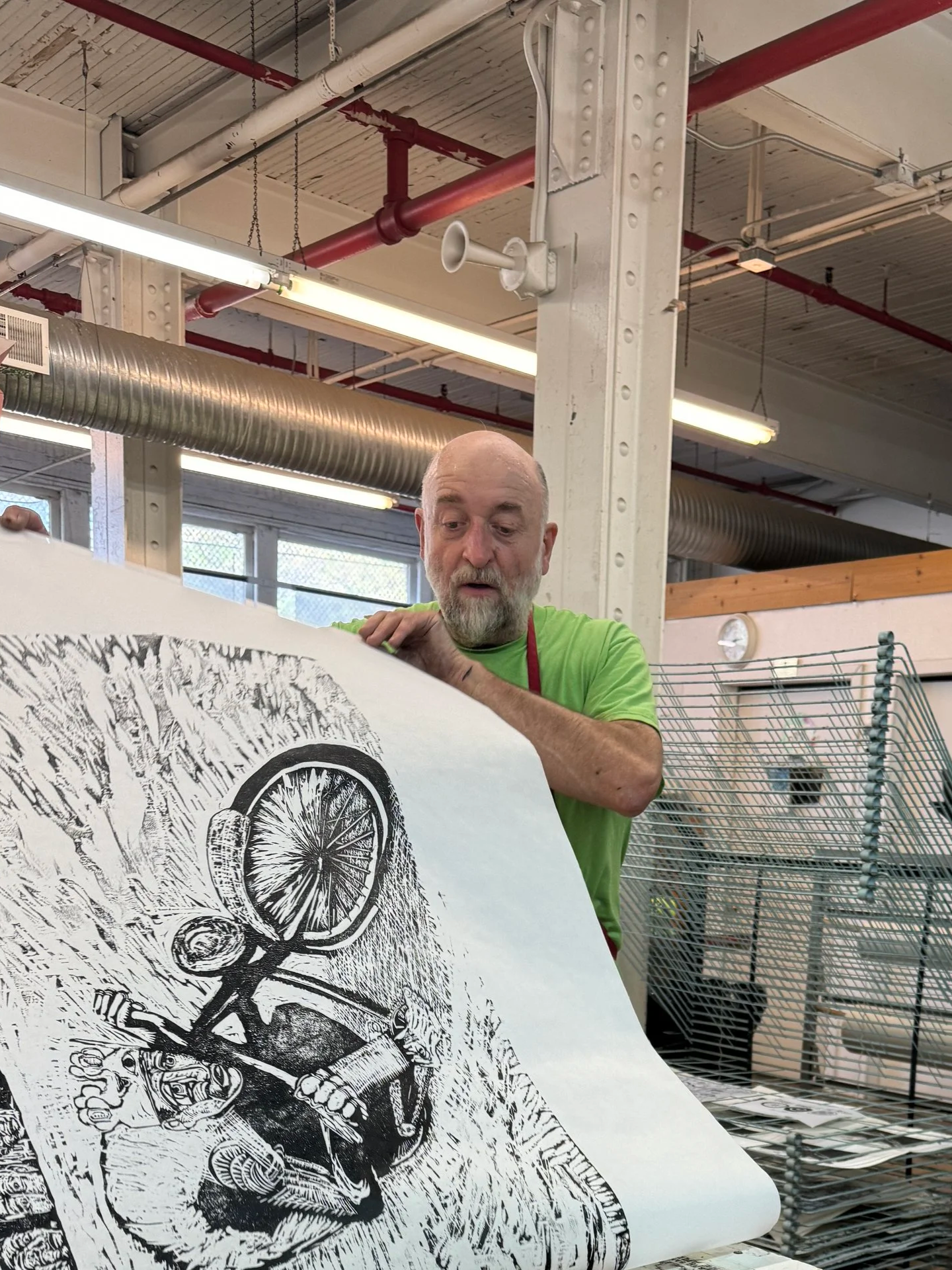 An older man with a bald head and beard examining a large black-and-white print of a detailed illustration of a motorcycle with a front wheel and engine, in an industrial setting with fluorescent lights and red pipes.