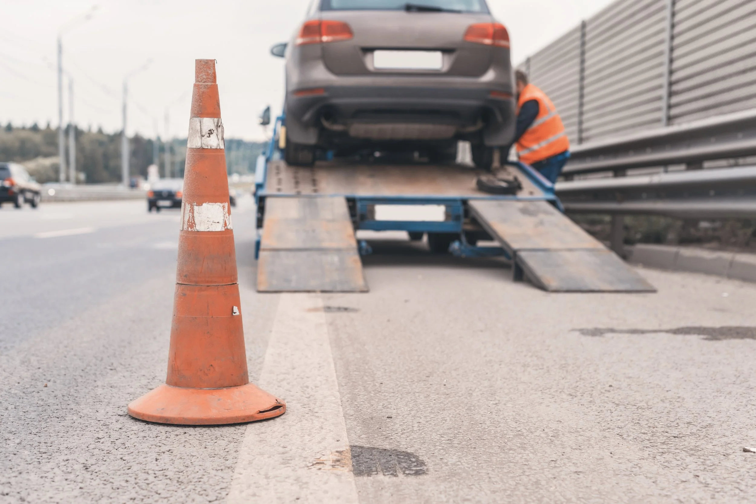 A roadside car being loaded onto a tow truck with an orange traffic cone in the foreground on a busy highway.