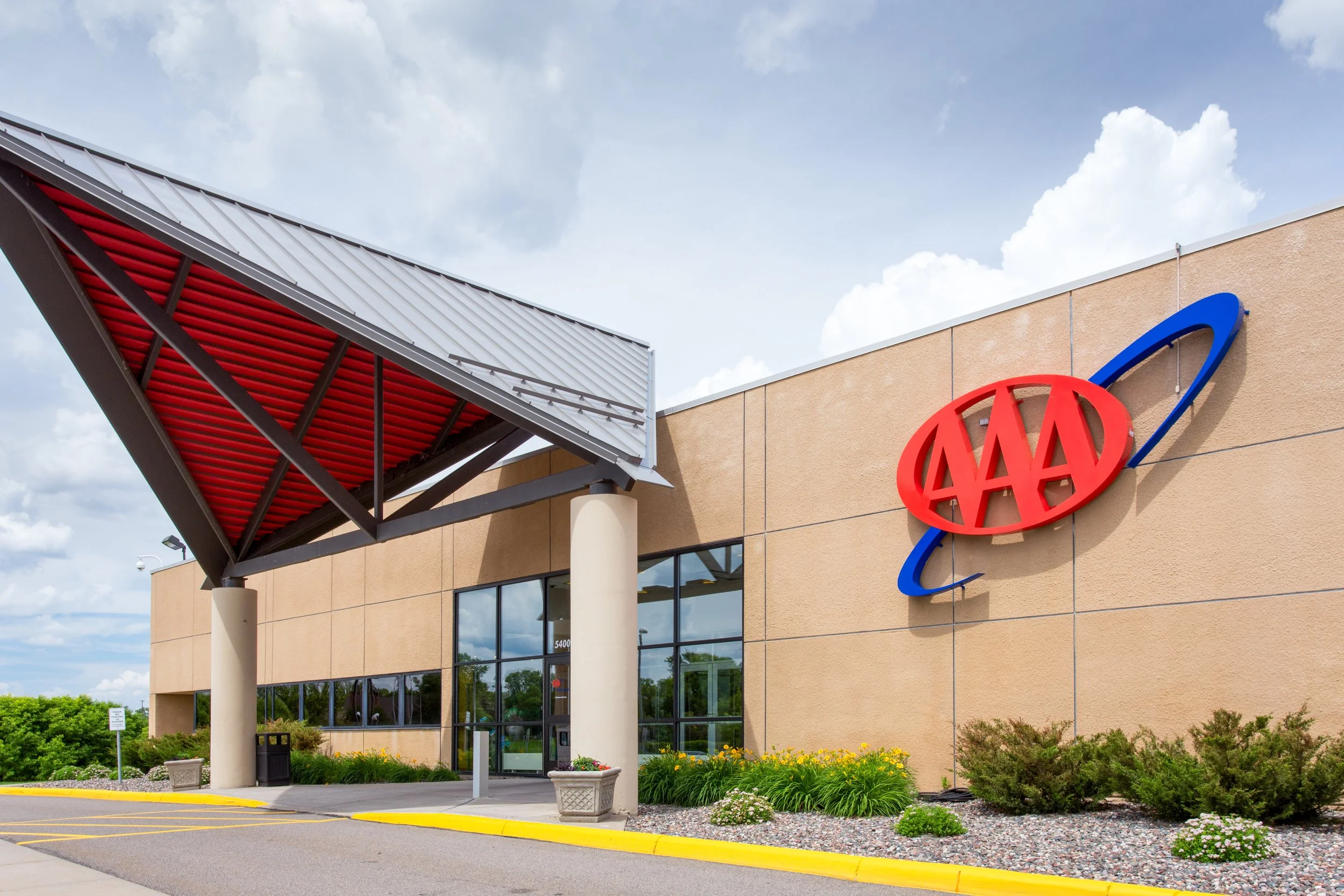 Exterior of AAA office building with a large AAA logo on the wall, a modern awning with red underside, and landscaped bushes and flowers in front.