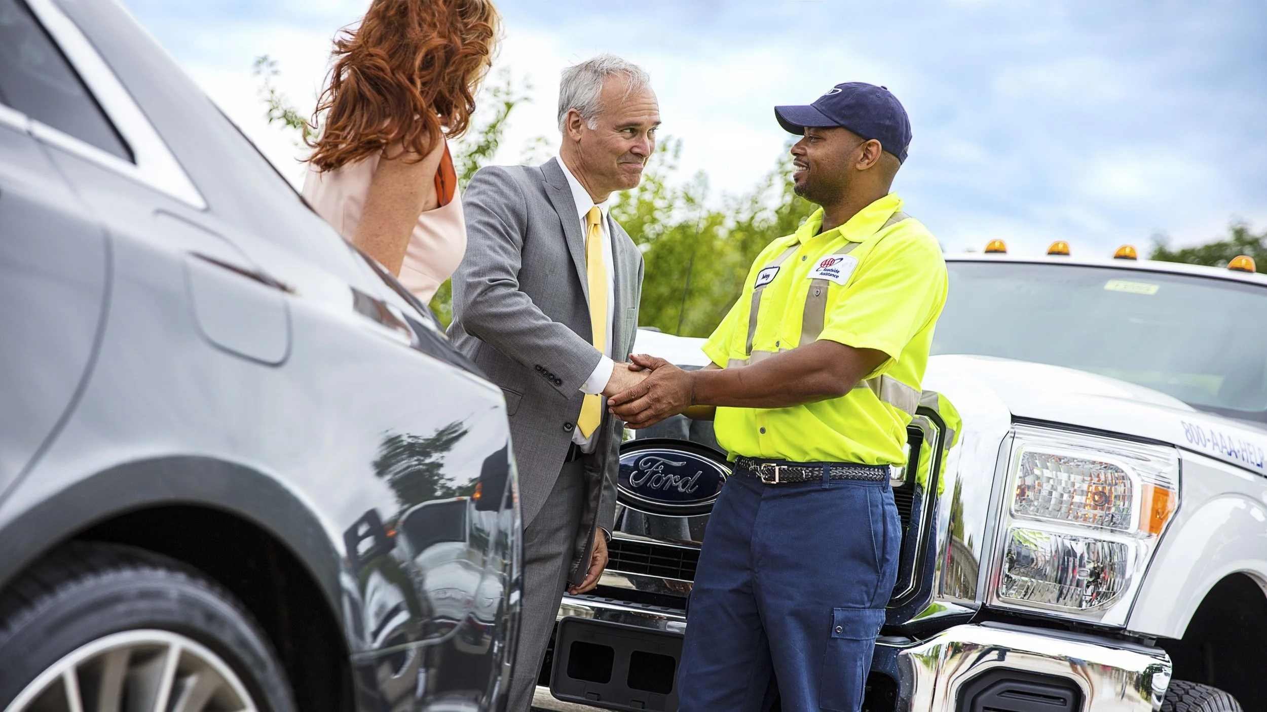 A man in a suit shaking hands with a tow truck worker beside a white tow truck, with a woman partially visible in the foreground, against a background of trees and cloudy sky.