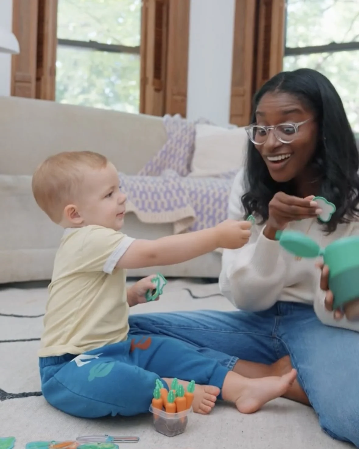 A woman with glasses and long black hair sitting on the floor, playing with a young boy in a living room with large windows and a beige couch. The boy is reaching out to the woman, holding a small toy. There are toys and a container of carrot juice on the floor.